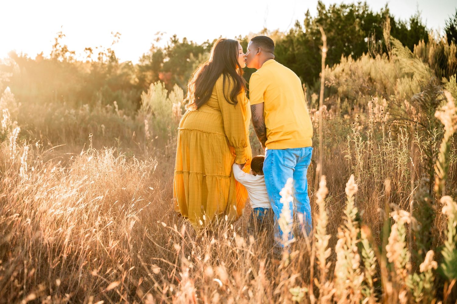 Couple kissing in sun-drenched golden field while young child stands between them at sunset.