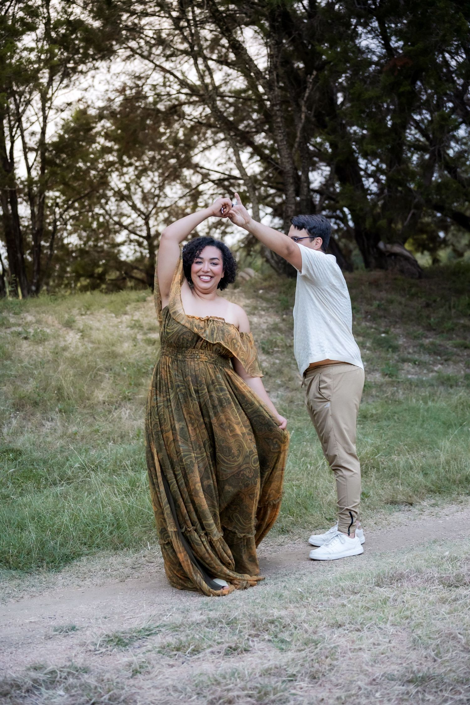 Couple dancing outdoors, man spinning pregnant woman in flowing green dress on a dirt path in a park.