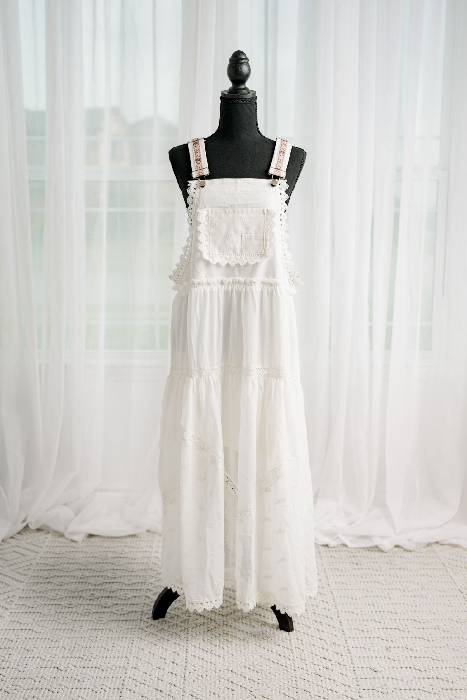 White eyelet lace overall-style maxi dress with decorative straps on black mannequin.