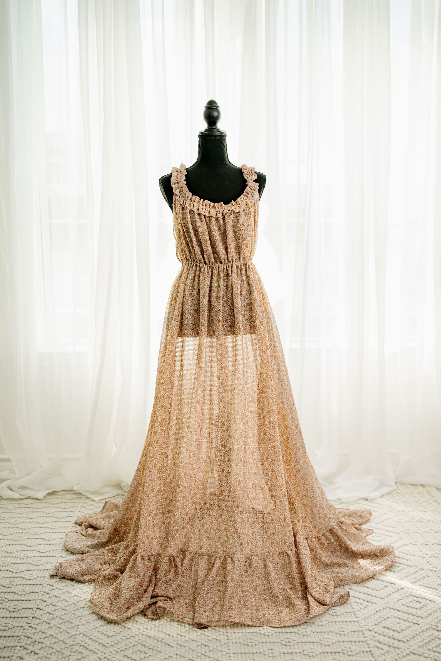 Elegant beige floral chiffon maxi dress on mannequin with ruffled neckline and flowing skirt displayed near sheer curtains.