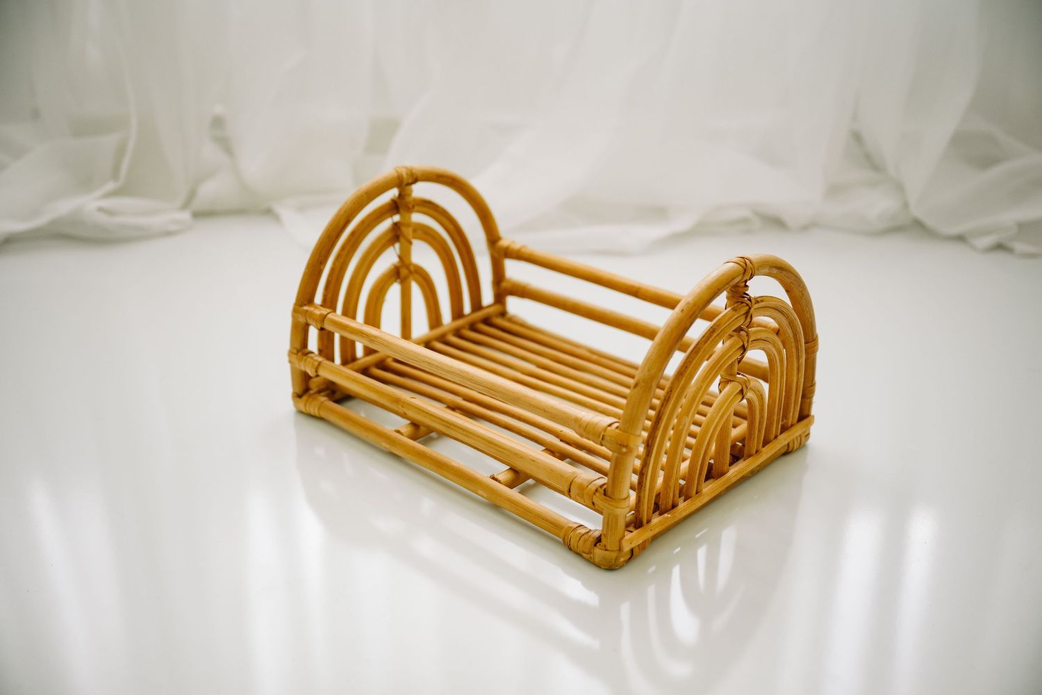 Miniature rattan arch-frame newborn photography prop bed on white reflective surface against sheer curtains.