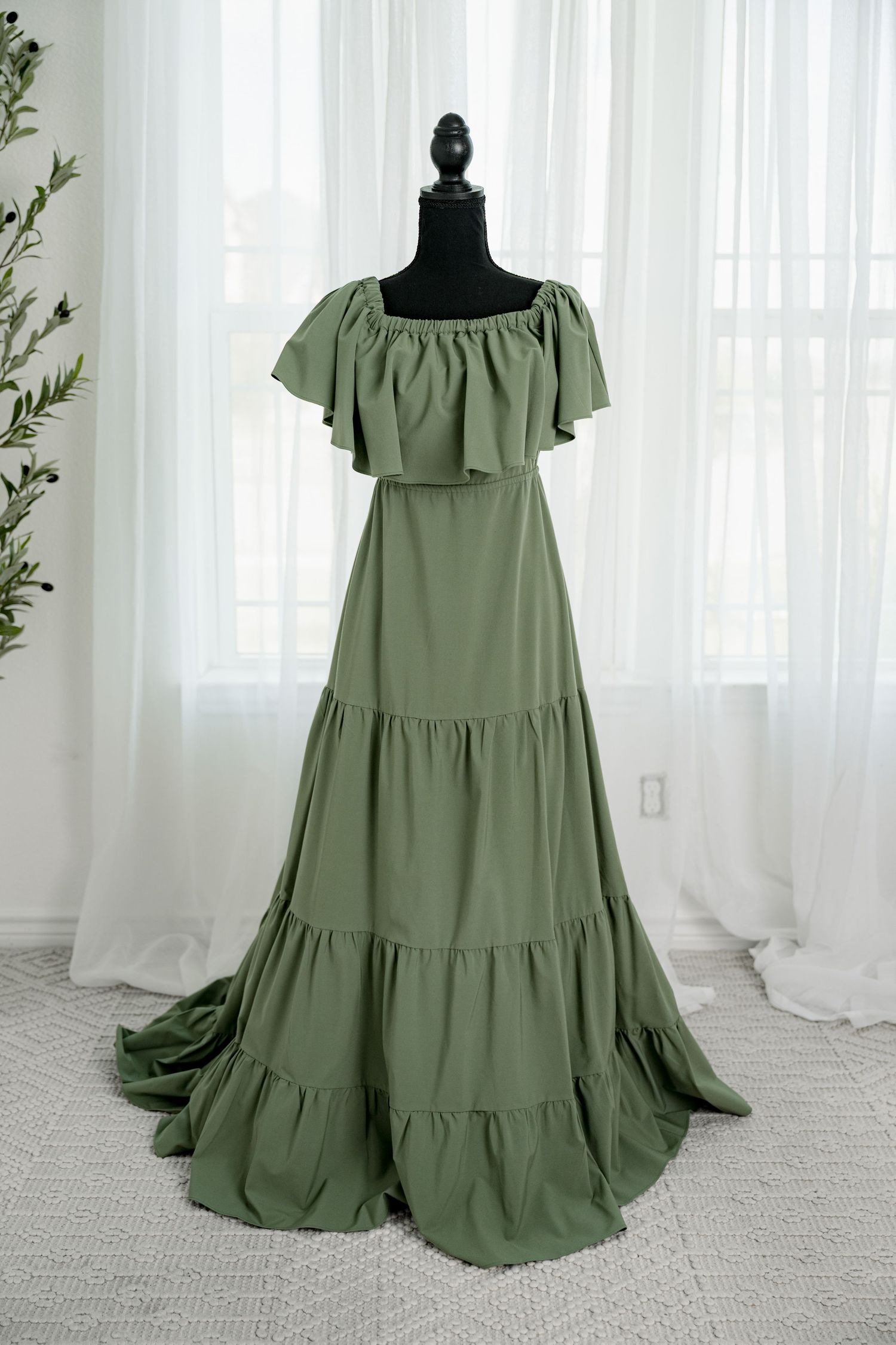 Sage green off-shoulder tiered maxi dress with ruffle neckline on a black mannequin against white curtains.