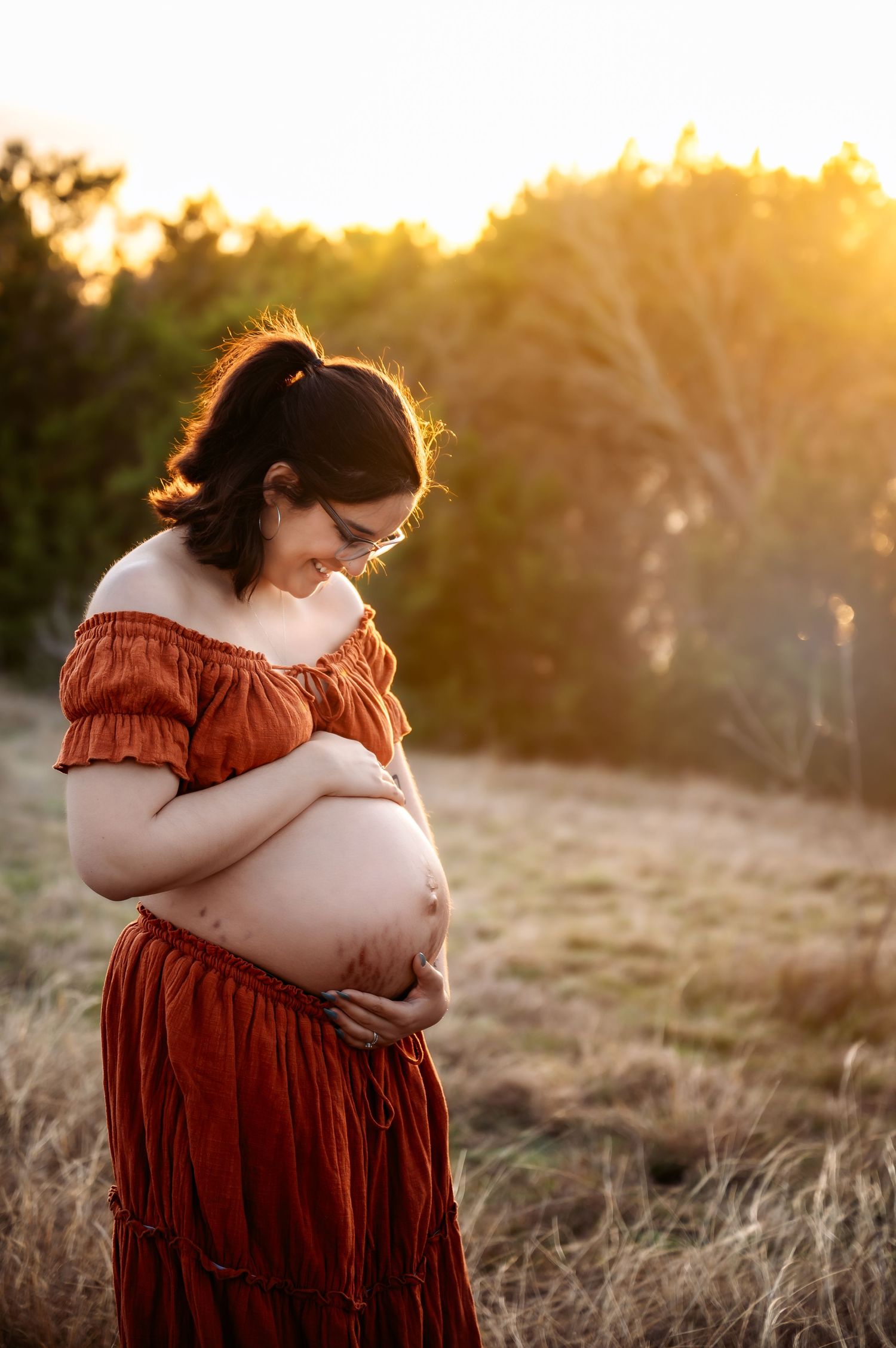 Pregnant woman in rust off-shoulder dress cradling belly at golden hour outdoors.