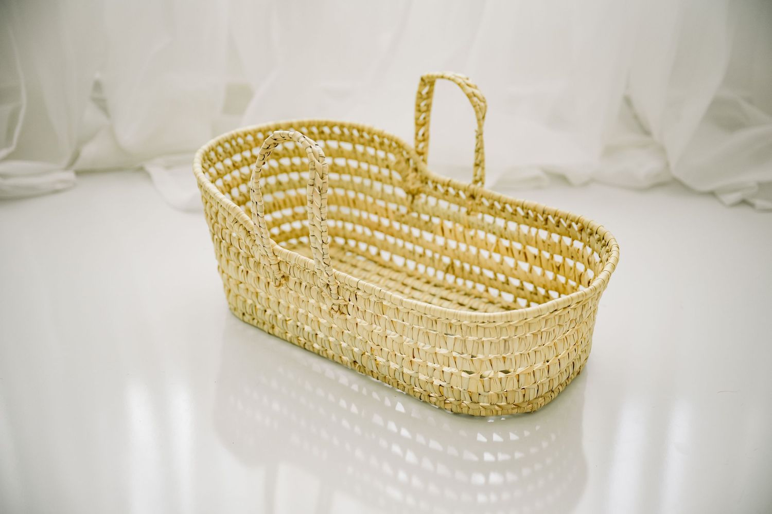 Woven natural seagrass Moses basket with handle on white reflective surface against sheer white curtains.