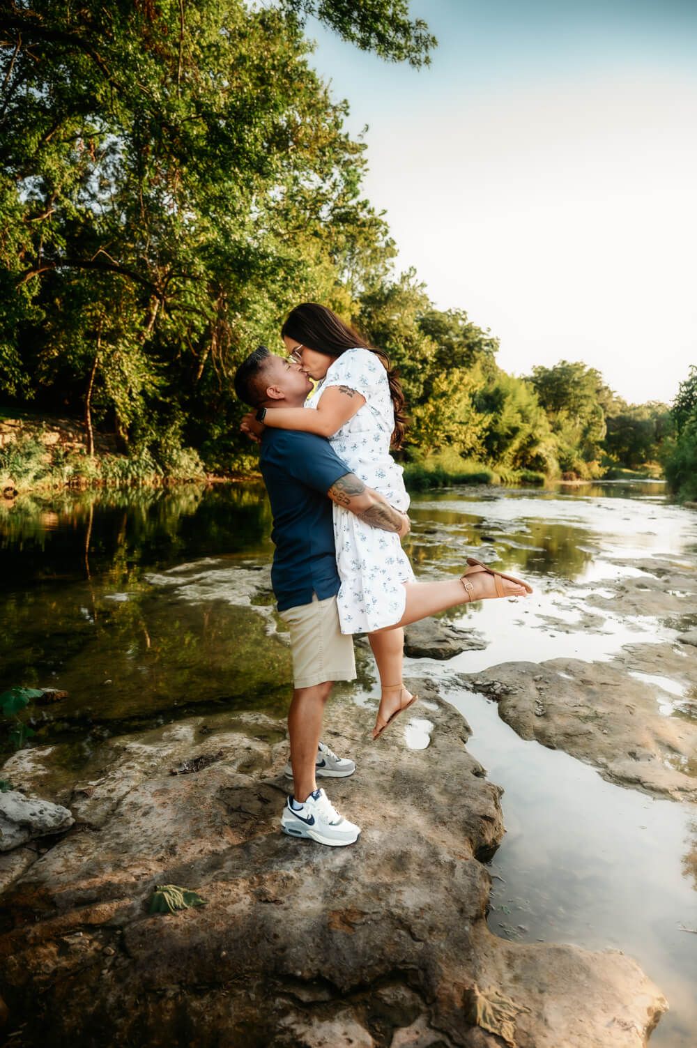 Couple embraces romantically on rocky riverbank during golden hour sunset.