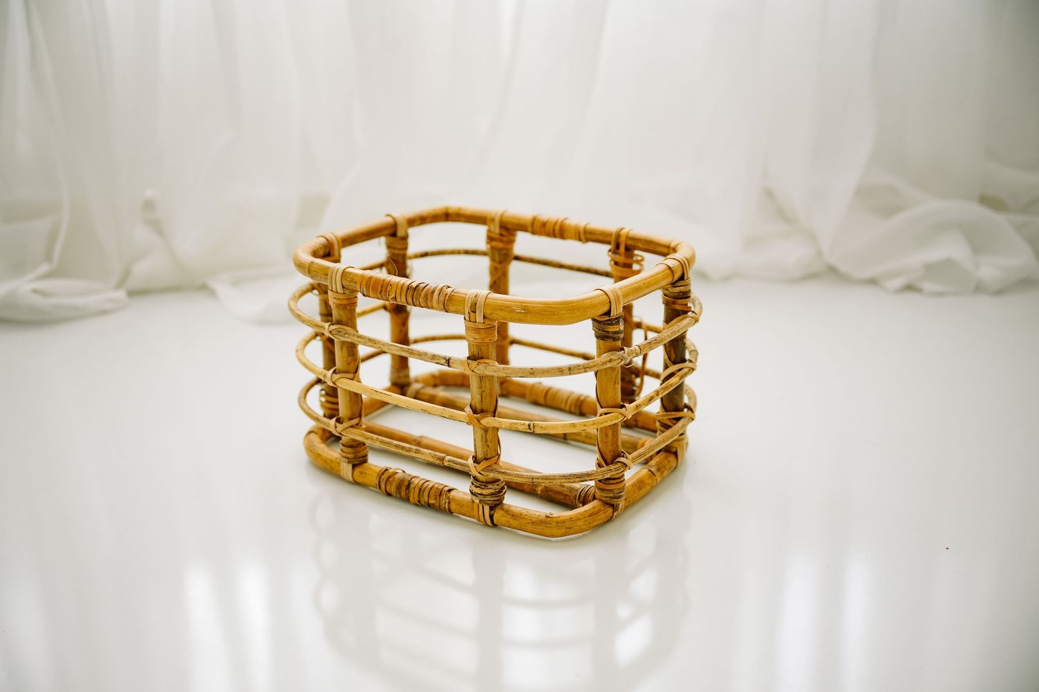 Square rattan bamboo storage basket on white reflective surface against sheer white curtains.