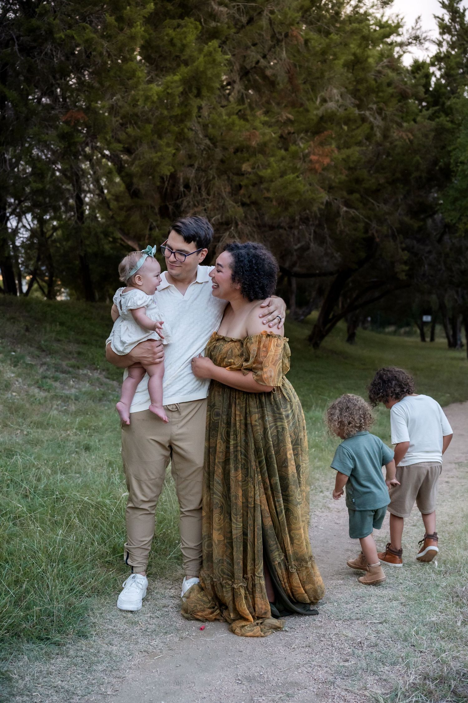 Family maternity photo in park, father holds baby while mother in green dress embraces him, toddler plays nearby.