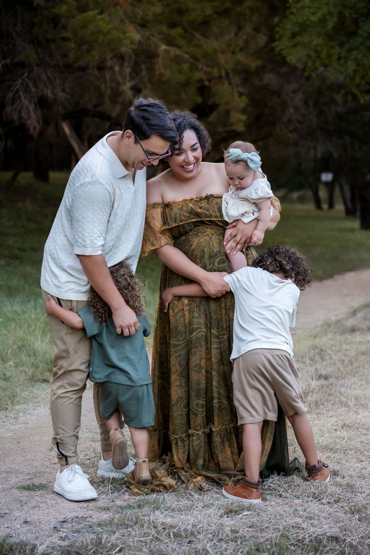 Pregnant woman in green dress laughs with family outdoors, husband and children touching her baby bump in a park.