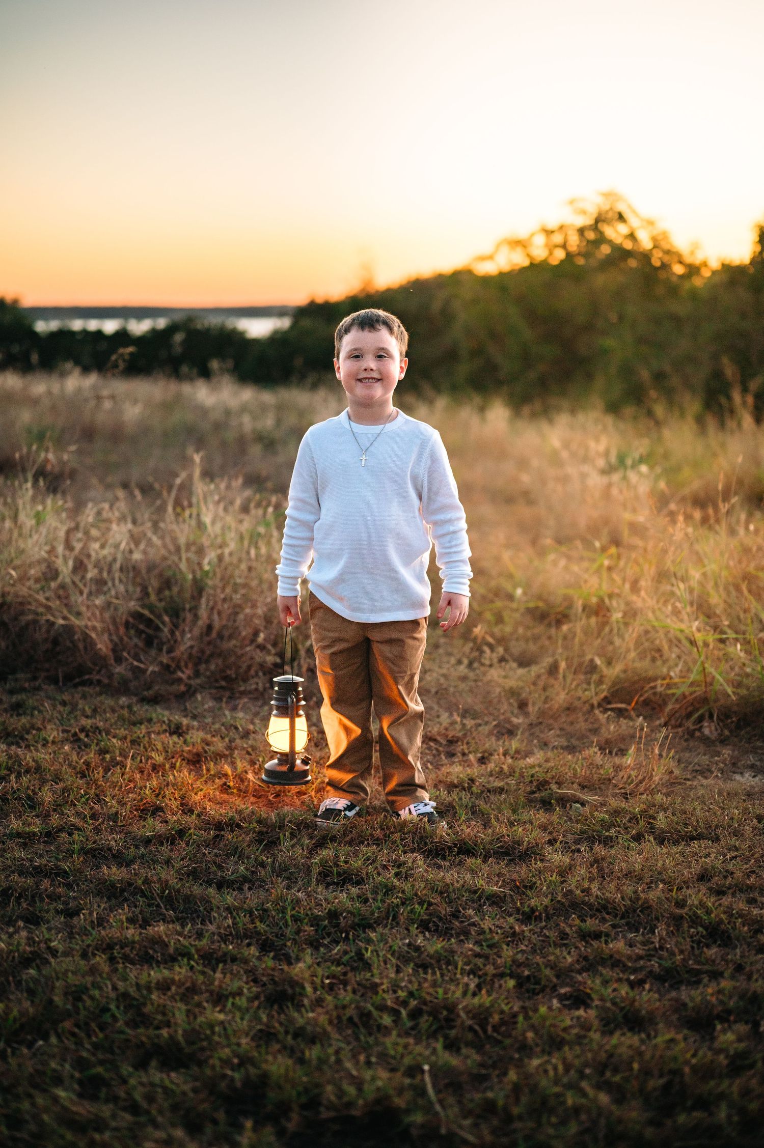 Smiling boy holding a vintage lantern while standing in a golden field at sunset.