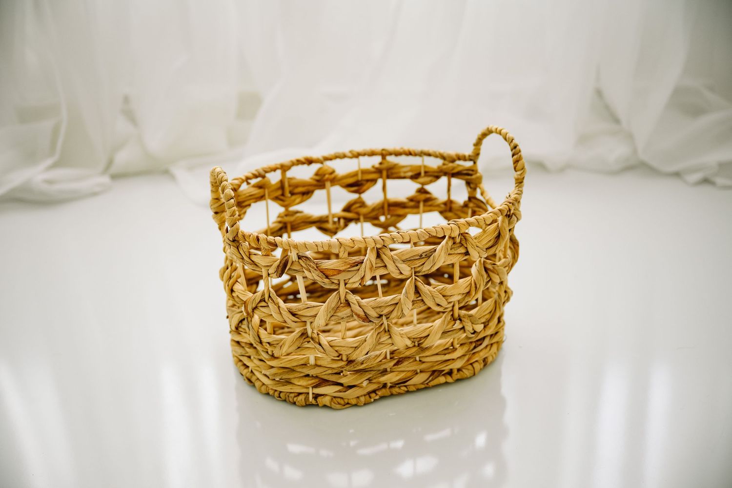 Round woven water hyacinth basket with side handles on white reflective surface against sheer curtains.