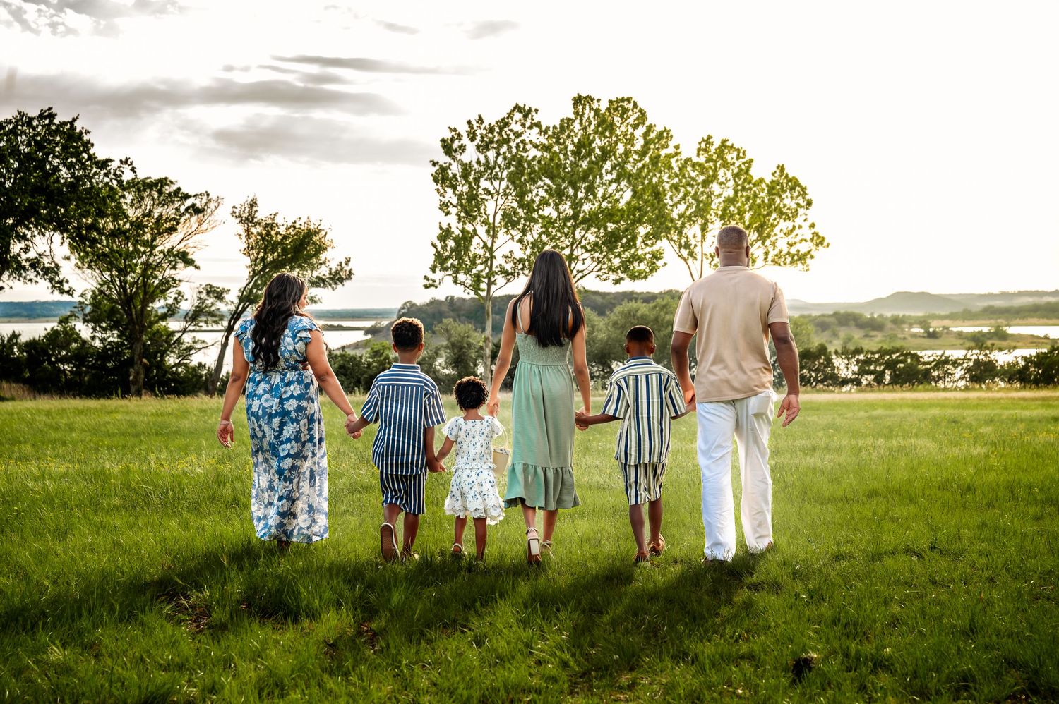 Family of six walking hand-in-hand through green meadow with scenic landscape in background.