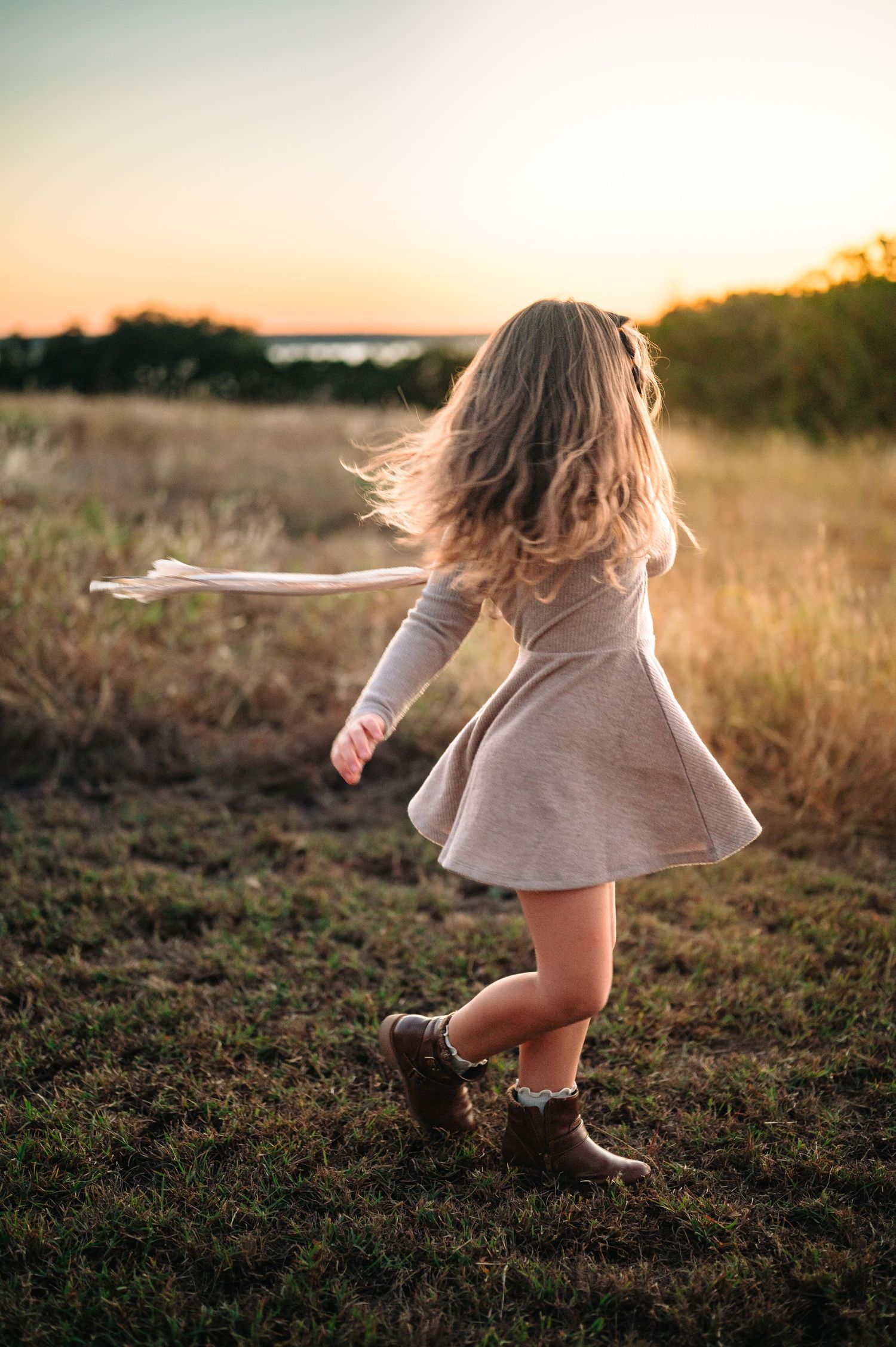 Young girl in a gray dress twirling in a field at sunset, hair flowing in the wind.