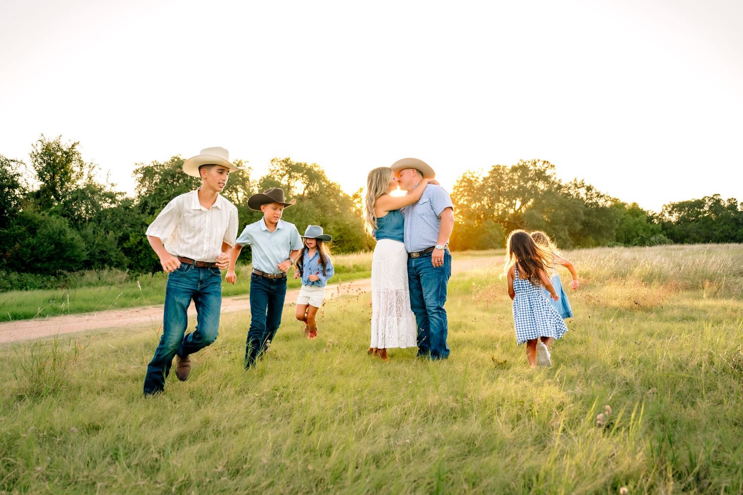 Family in cowboy hats kissing and playing in a sunlit green field at golden hour.