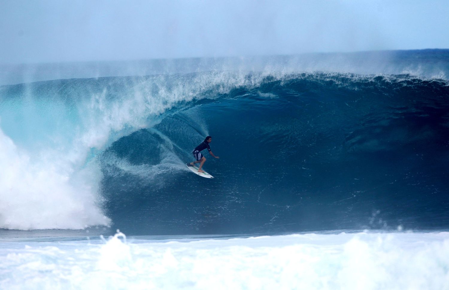 A surfer rides a massive blue wave at Pipeline in Hawaii as white spray erupts from the powerful ocean swell.