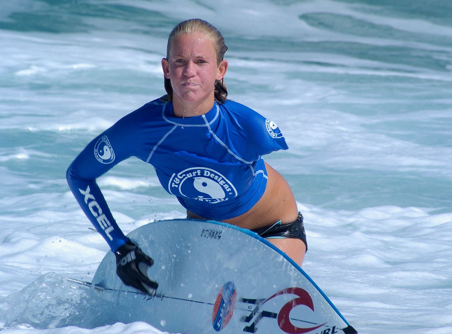 A surfer in a blue rash guard rides a white surfboard on turquoise ocean waves.