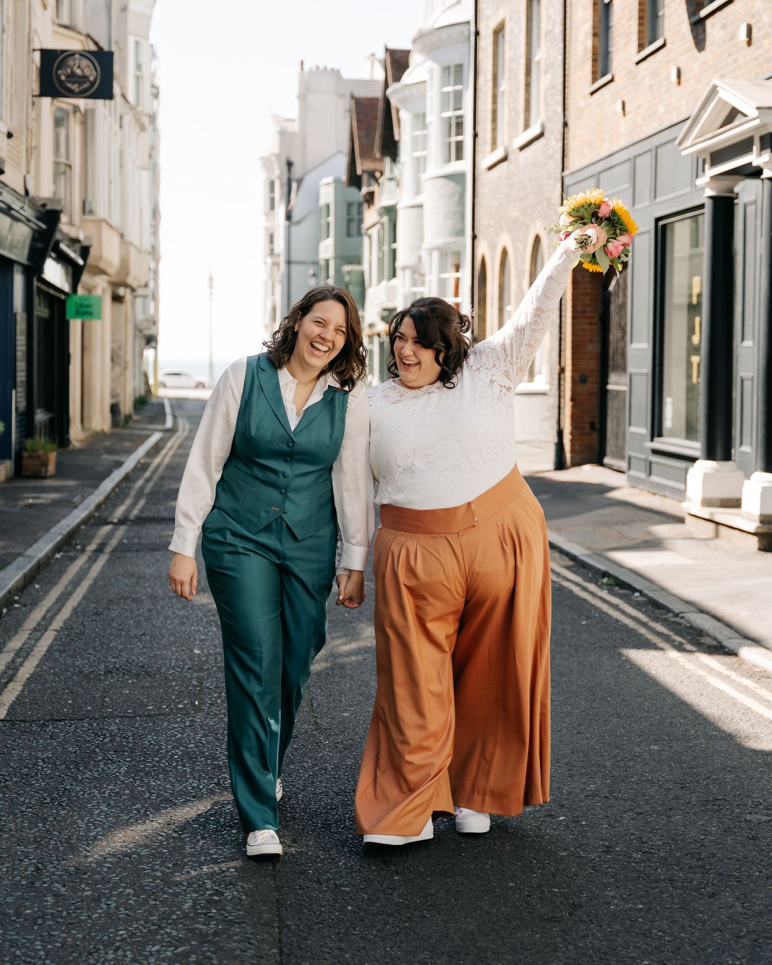 Two brides walk joyfully down a sunny street, one in green suit, one in orange trousers holding sunflowers.