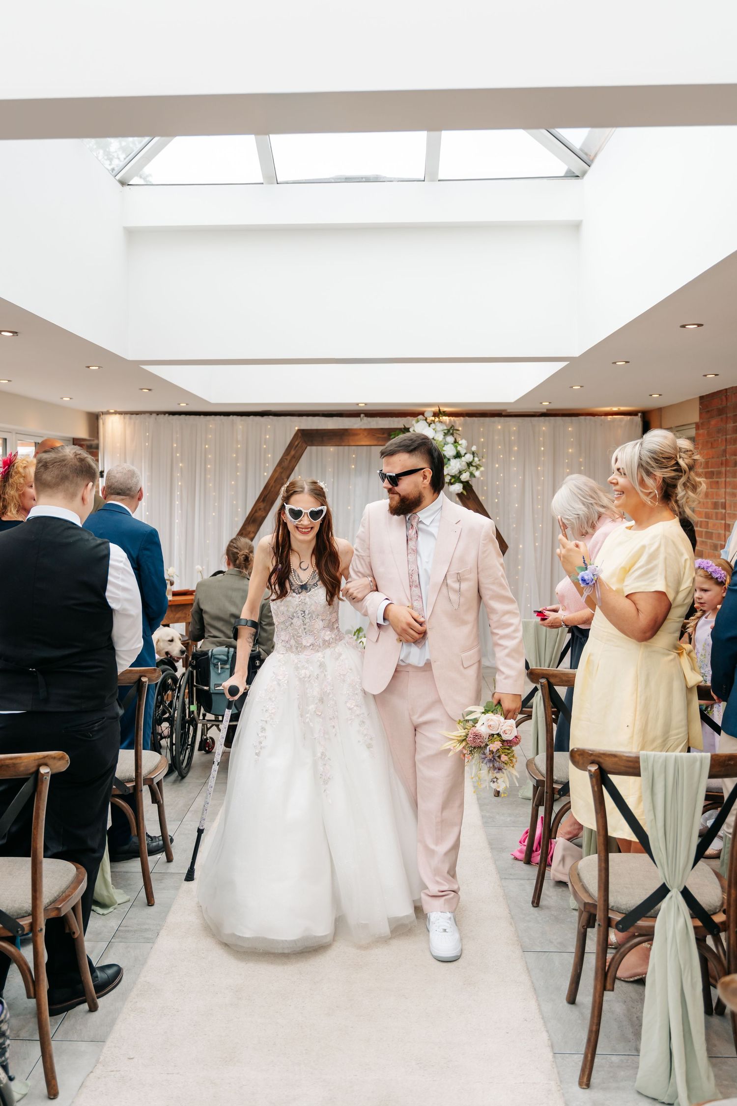 Bride and groom walk down the aisle in matching pink outfits and sunglasses at a modern wedding venue.