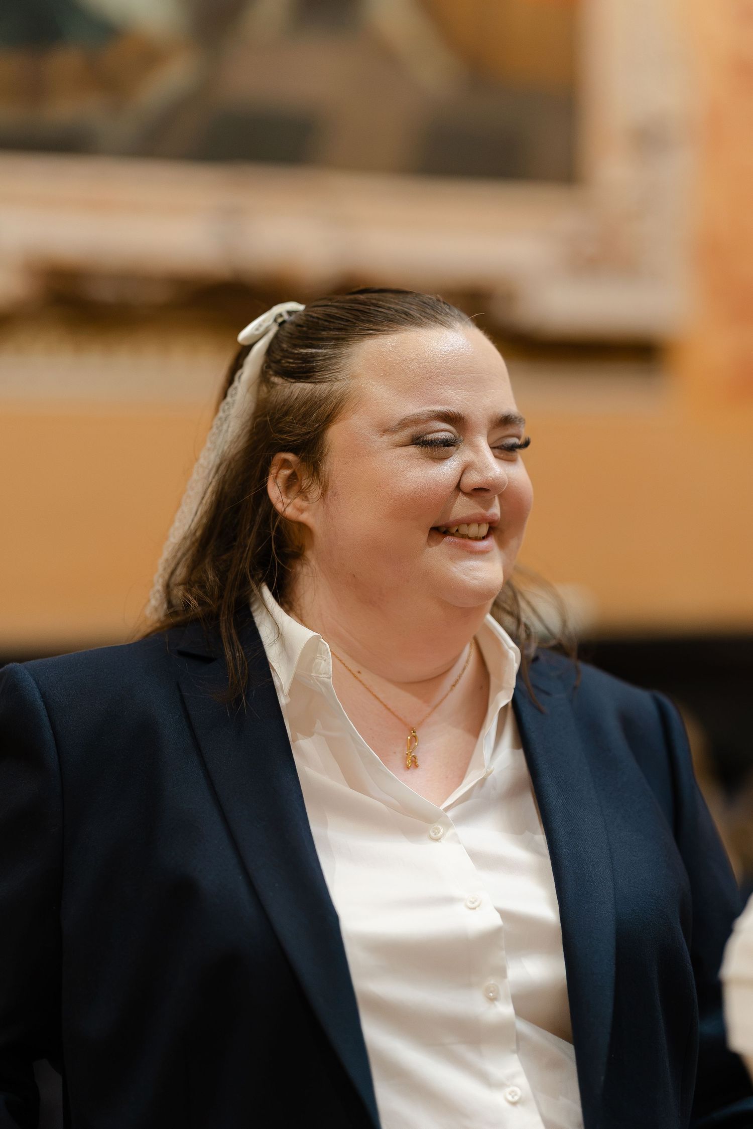 Woman in navy blazer and white shirt smiling, wearing gold necklace, hair up with white bow accessory.
