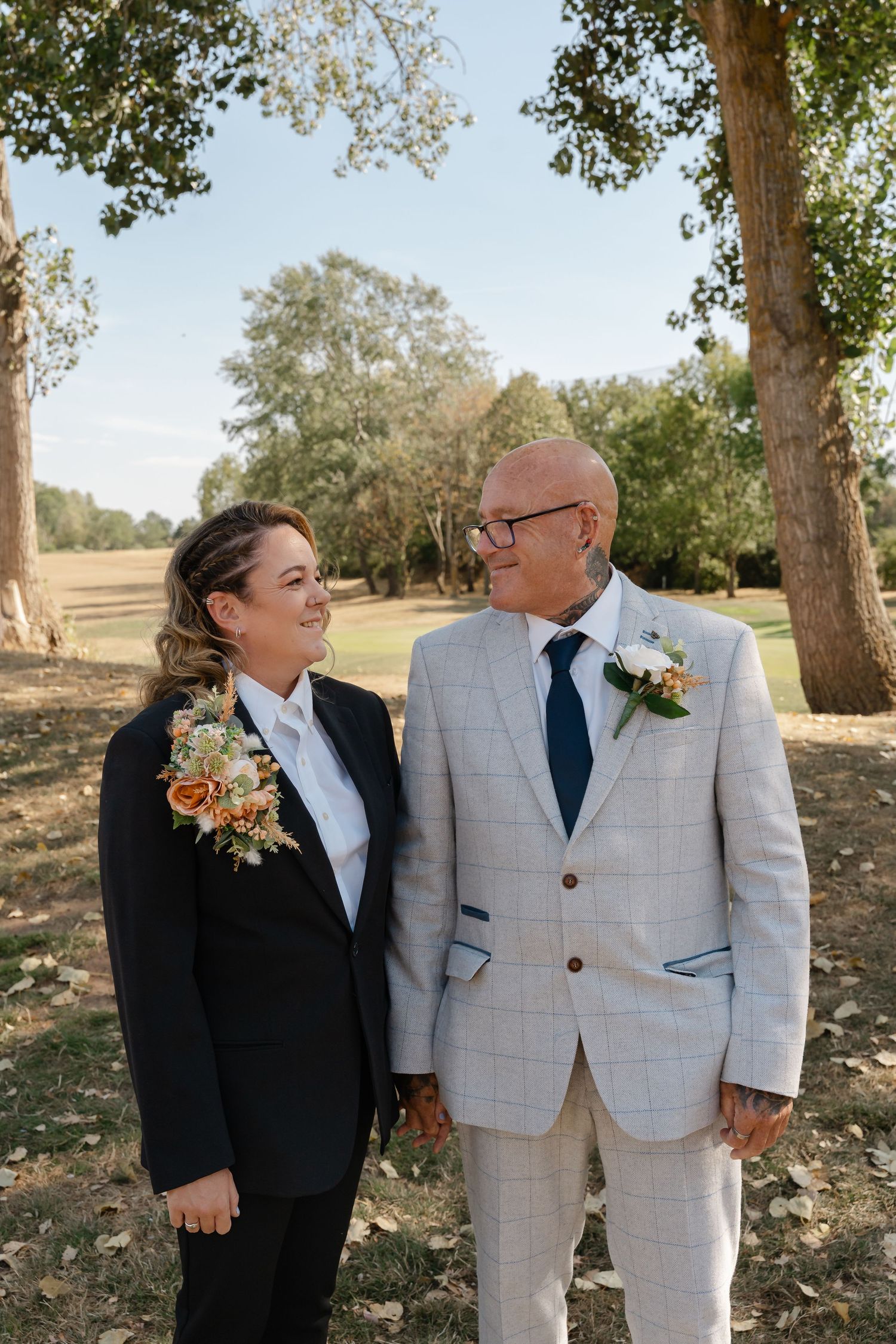 Couple in formal wedding attire with boutonnieres smiling at each other outdoors under trees on a sunny day.