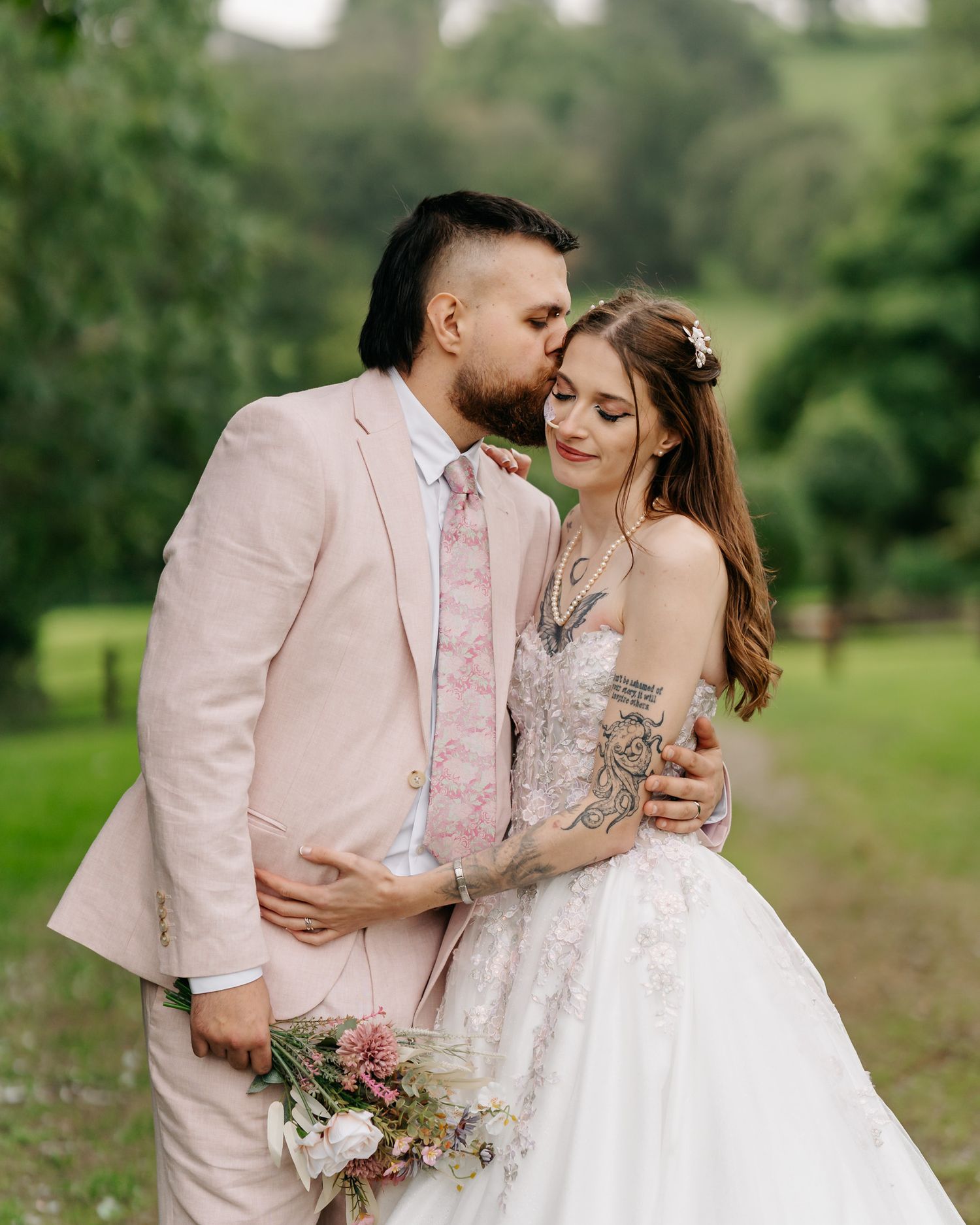 Couple in wedding attire, groom in pink suit kissing bride in lace gown, holding floral bouquet outdoors.
