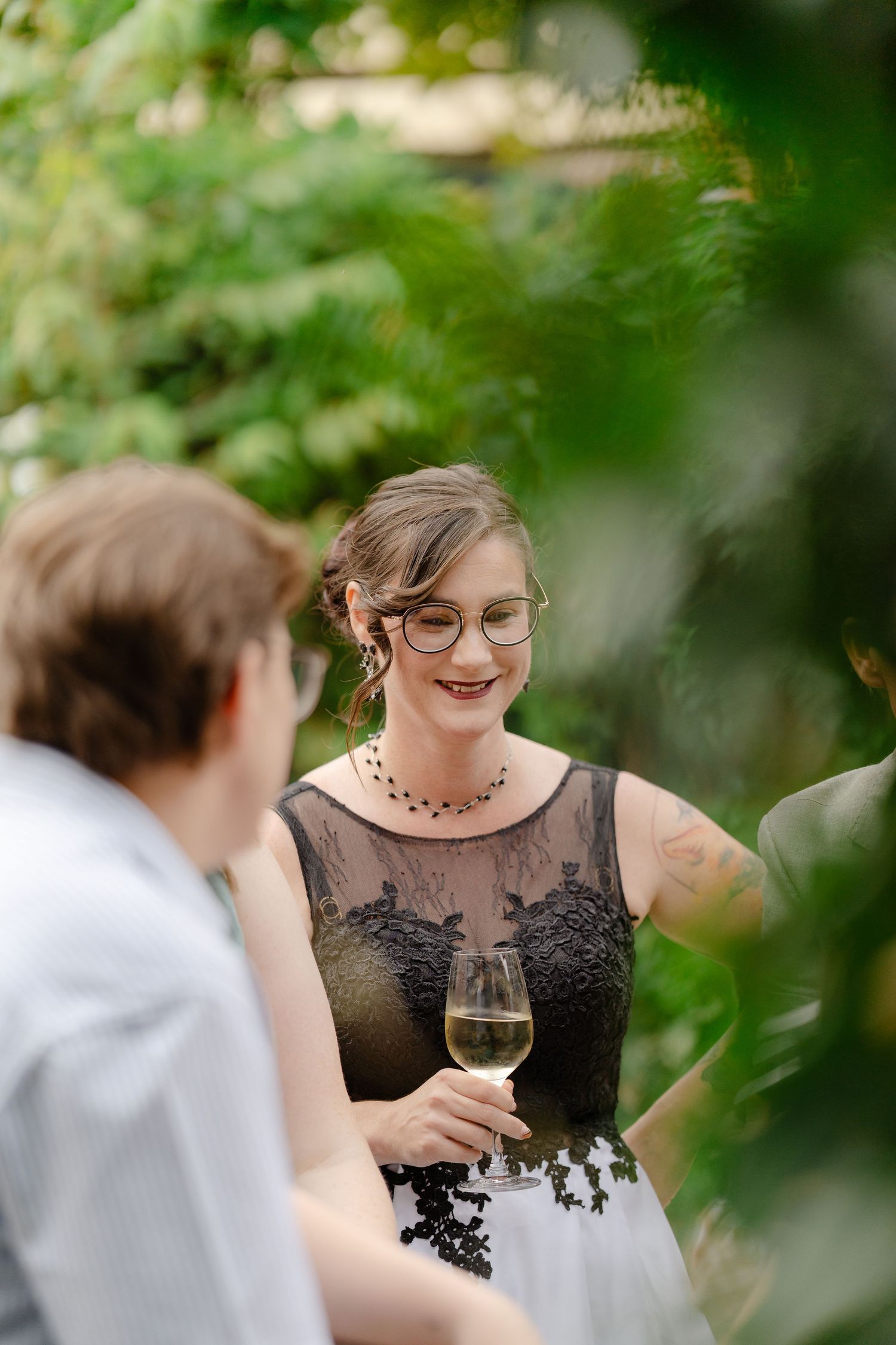 Woman in black lace dress holding white wine glass, smiling outdoors among green foliage at a garden event.