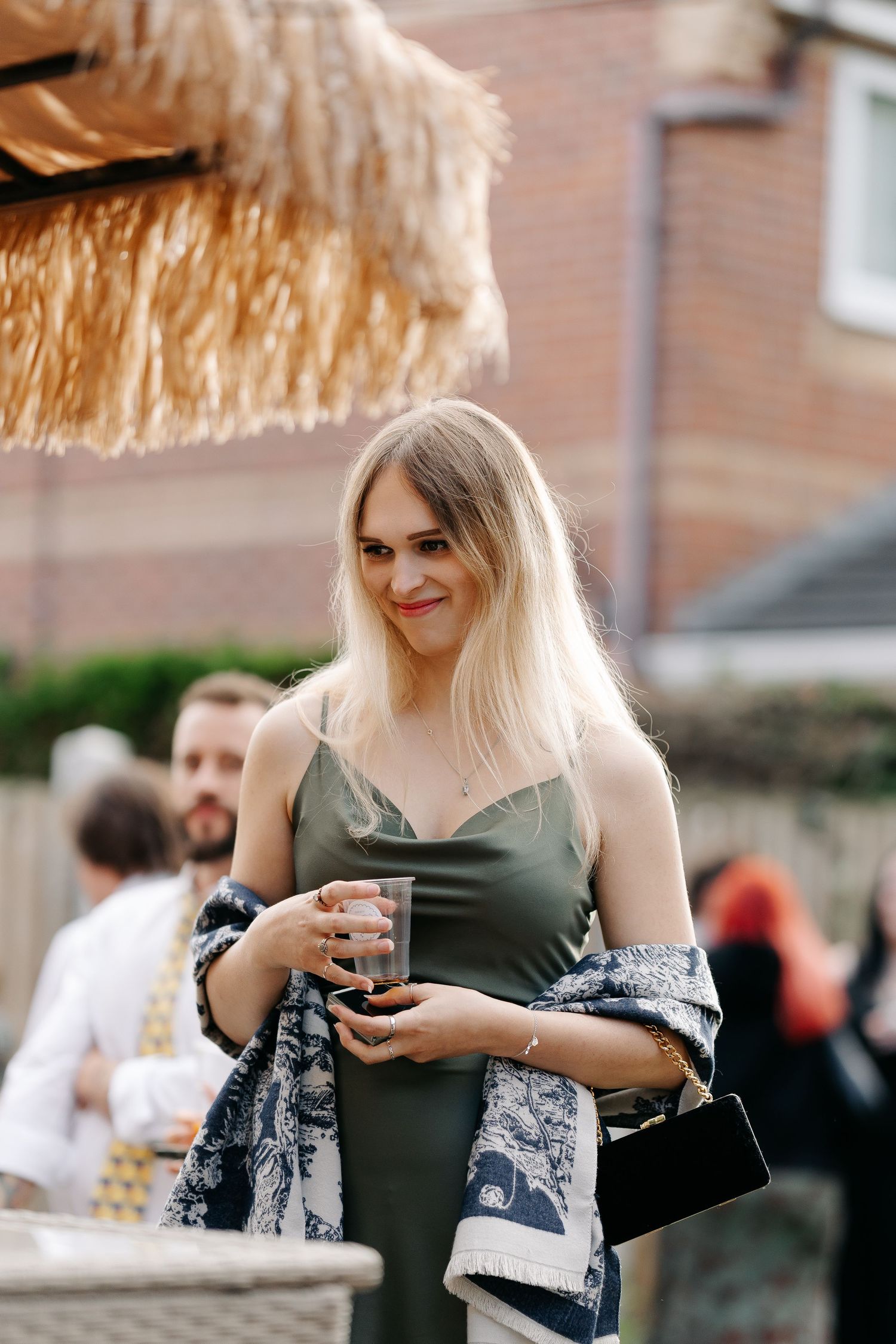 Blonde woman in olive slip dress holding a drink at an outdoor party, wearing a patterned kimono and carrying a black clutch.