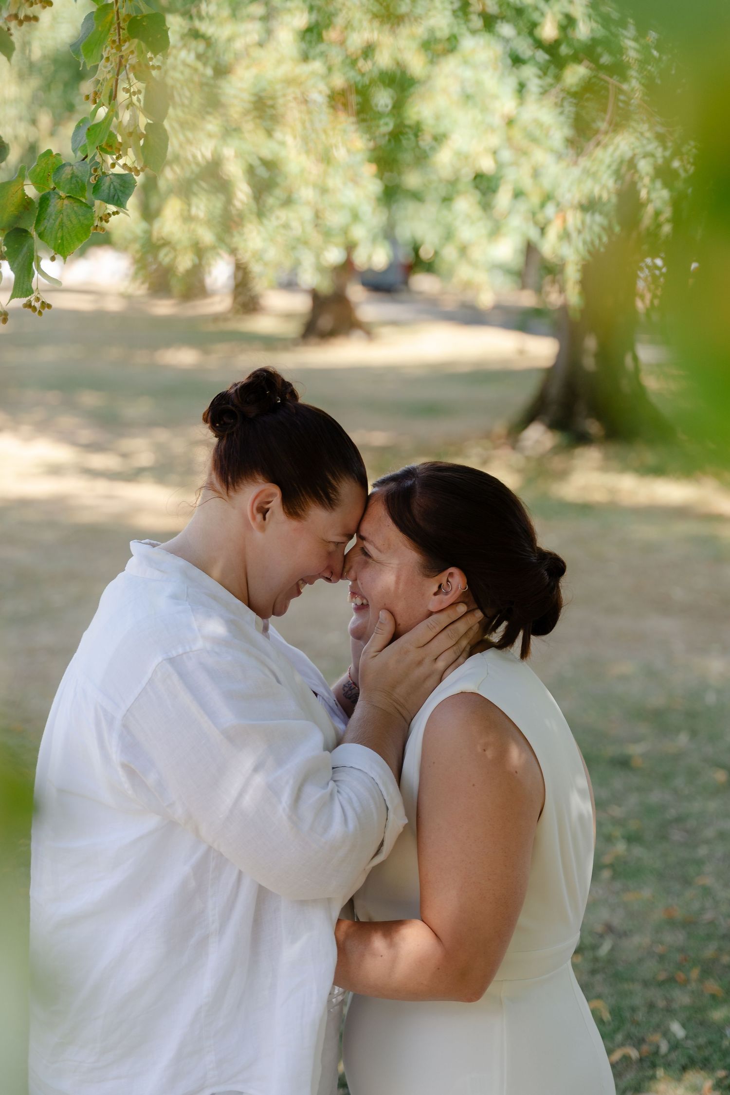 Two women in white outfits share a tender forehead touch in a sunlit park setting.