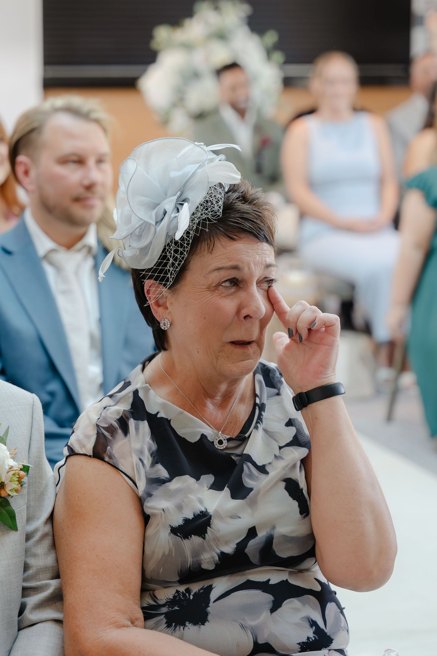 Emotional woman wiping tears at a wedding ceremony, wearing a floral dress and elegant fascinator hat.