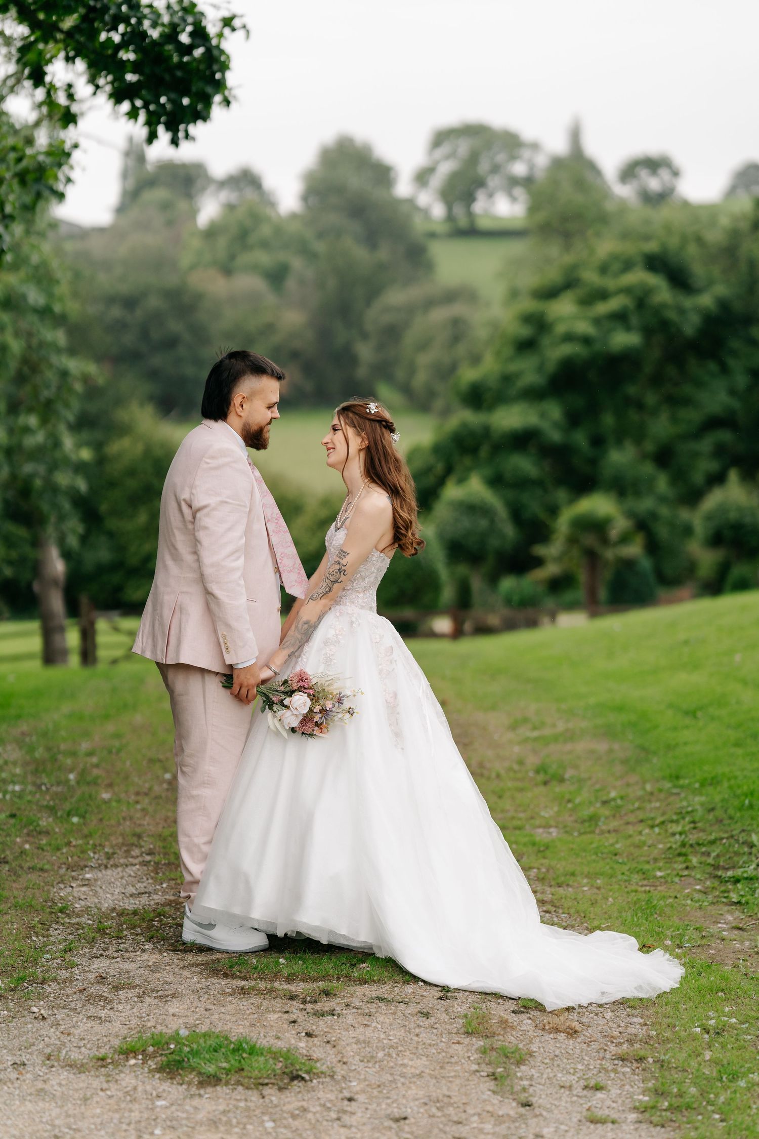 Bride and groom holding hands in a lush green park, bride in white gown with bouquet, groom in light pink suit.
