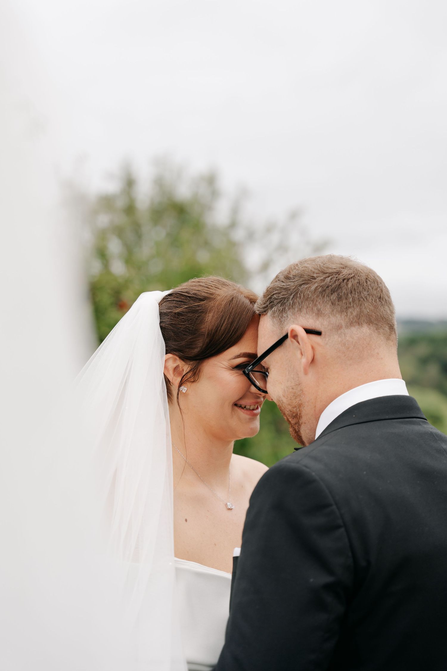 A smiling bride in a white veil and groom in a black suit touch foreheads tenderly during their wedding day.