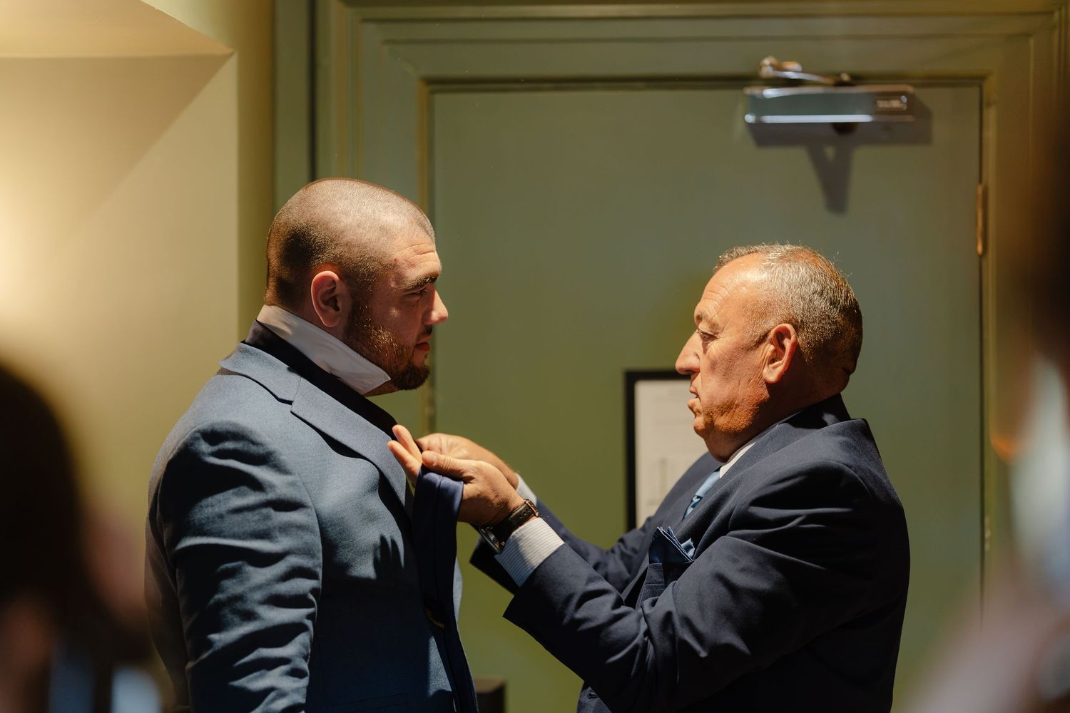 Two men in suits adjusting a tie near a door in a formal indoor setting.