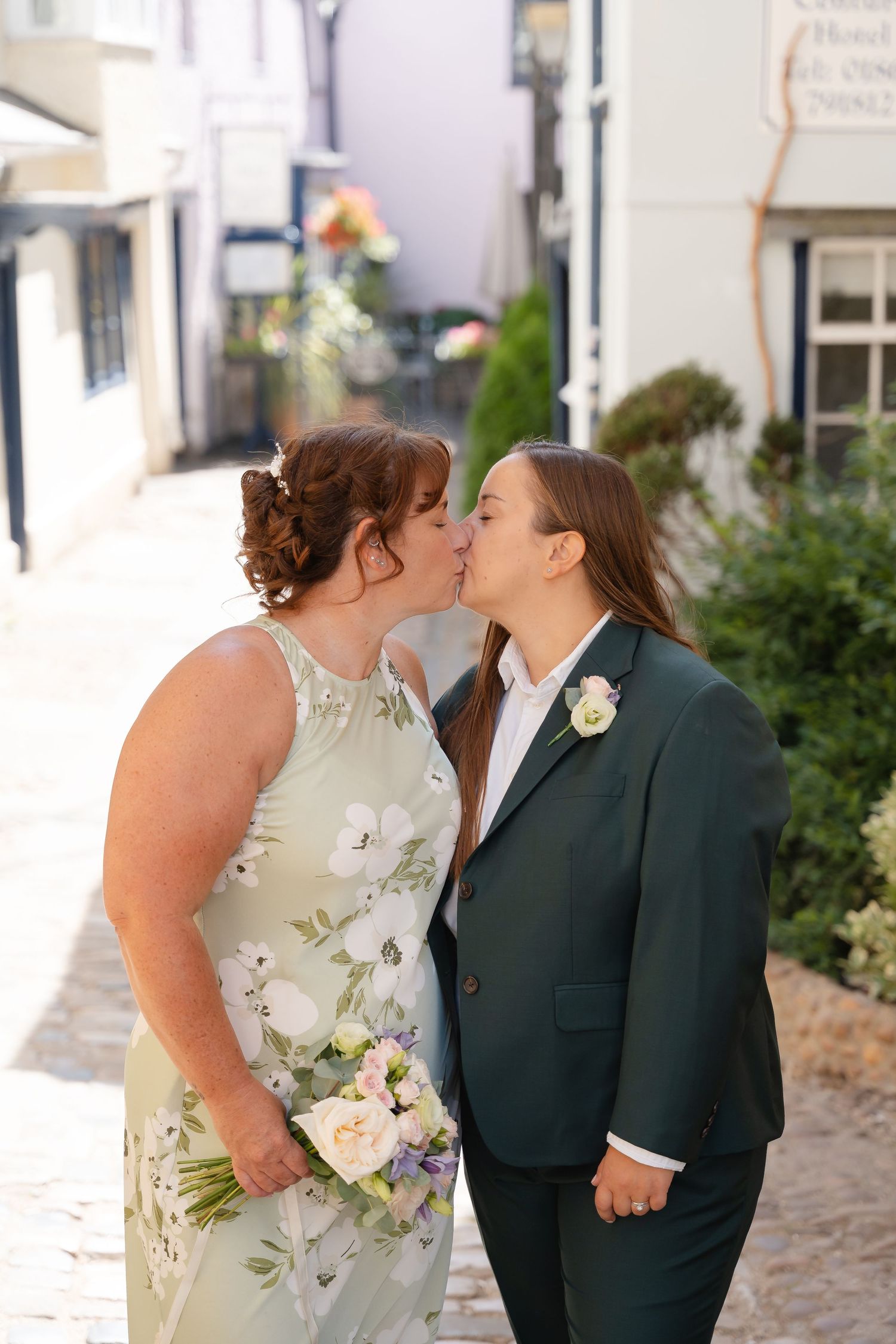 Two brides sharing a kiss on a cobblestone street, one in a floral dress holding a bouquet, the other in a green suit.
