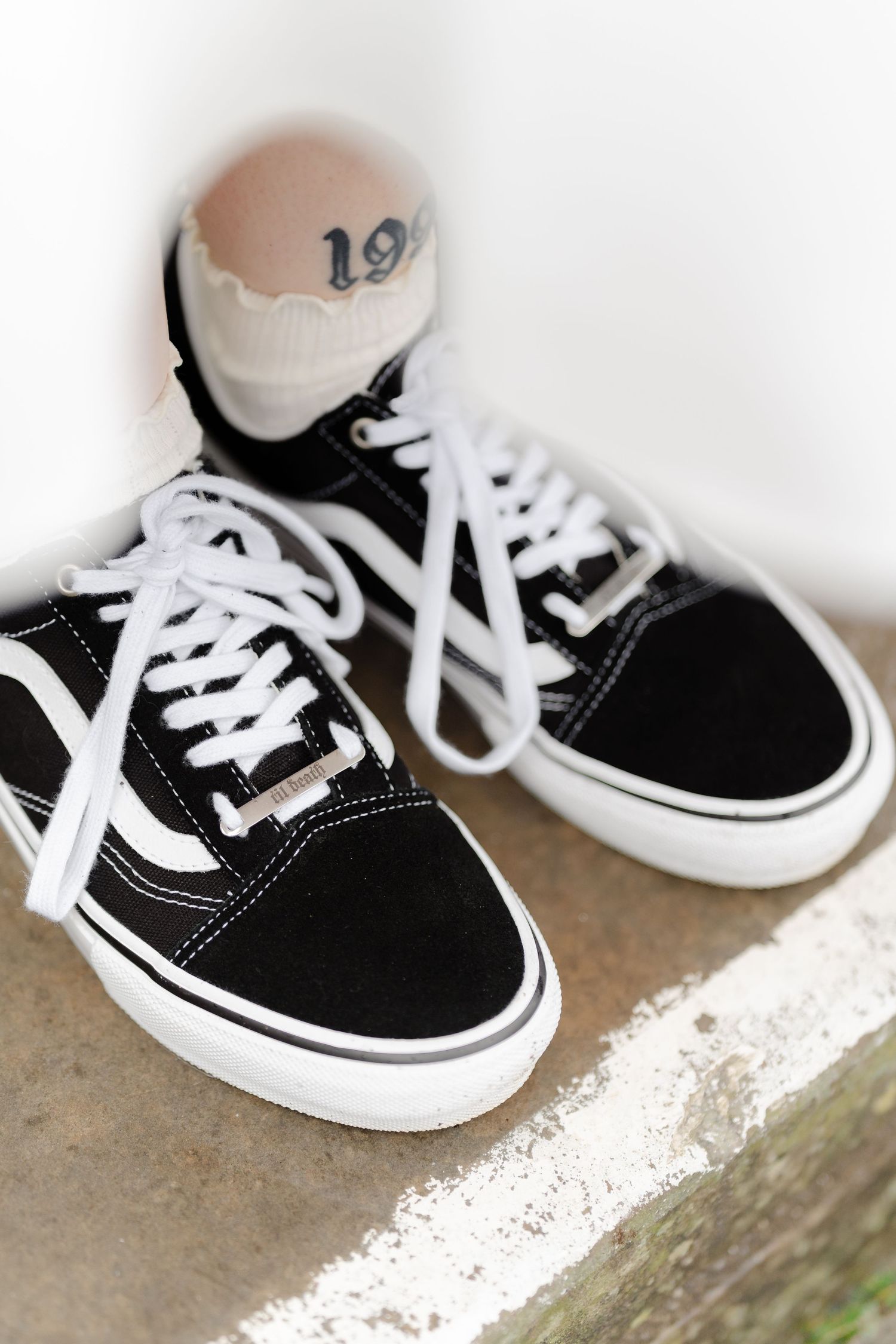 Black and white Vans Old Skool sneakers with white laces on a concrete step, with a tattooed ankle visible.