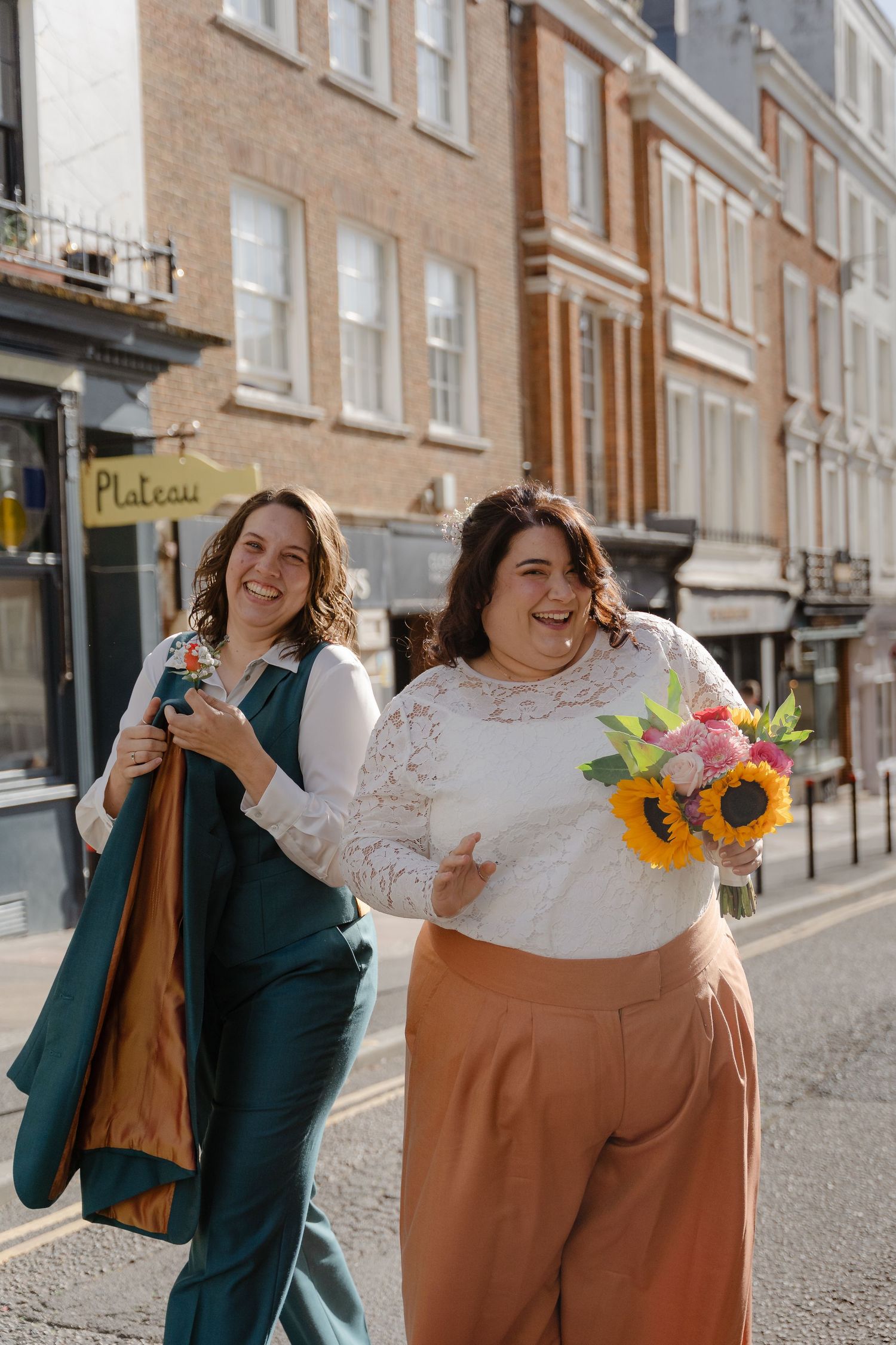 Two brides laughing on a sunny street, one holding sunflowers, wearing lace top and teal suit outside brick buildings.