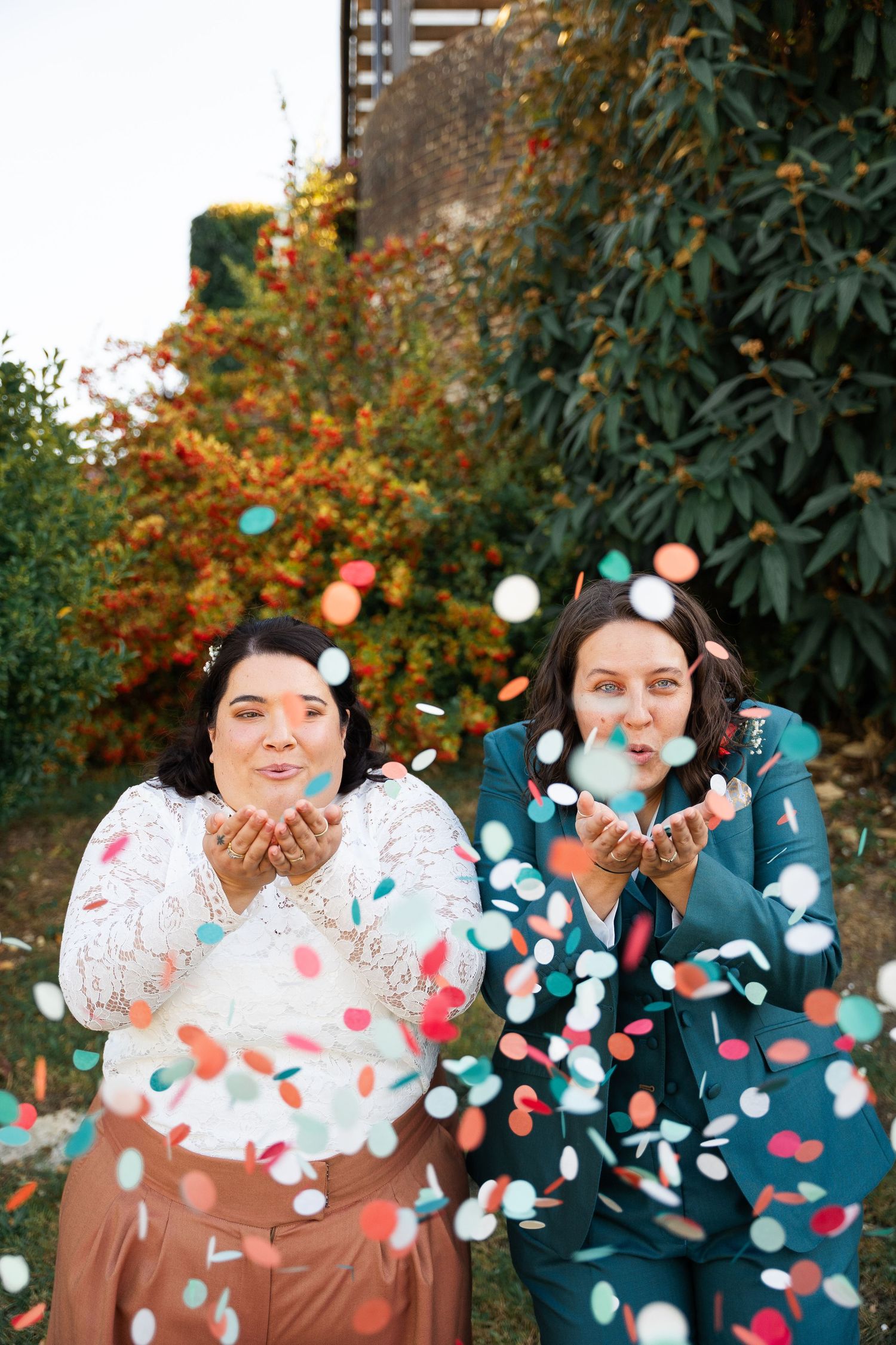 Two women in festive attire blow colorful confetti from their hands outdoors, celebrating joyfully.