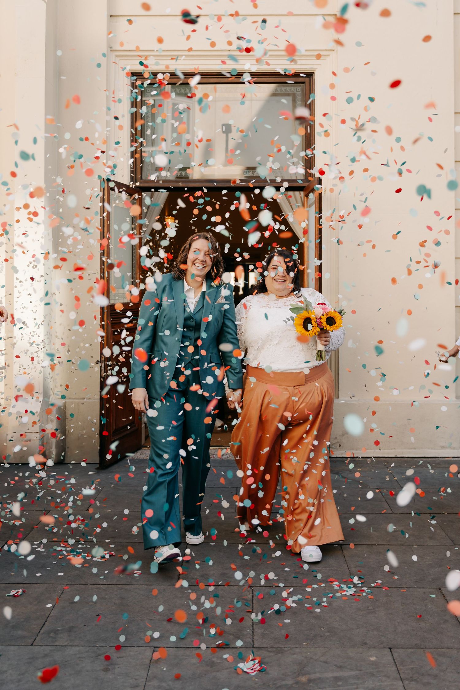 Two brides celebrate with colorful confetti outside a building, one in a teal suit and one in a copper skirt holding sunflowers.