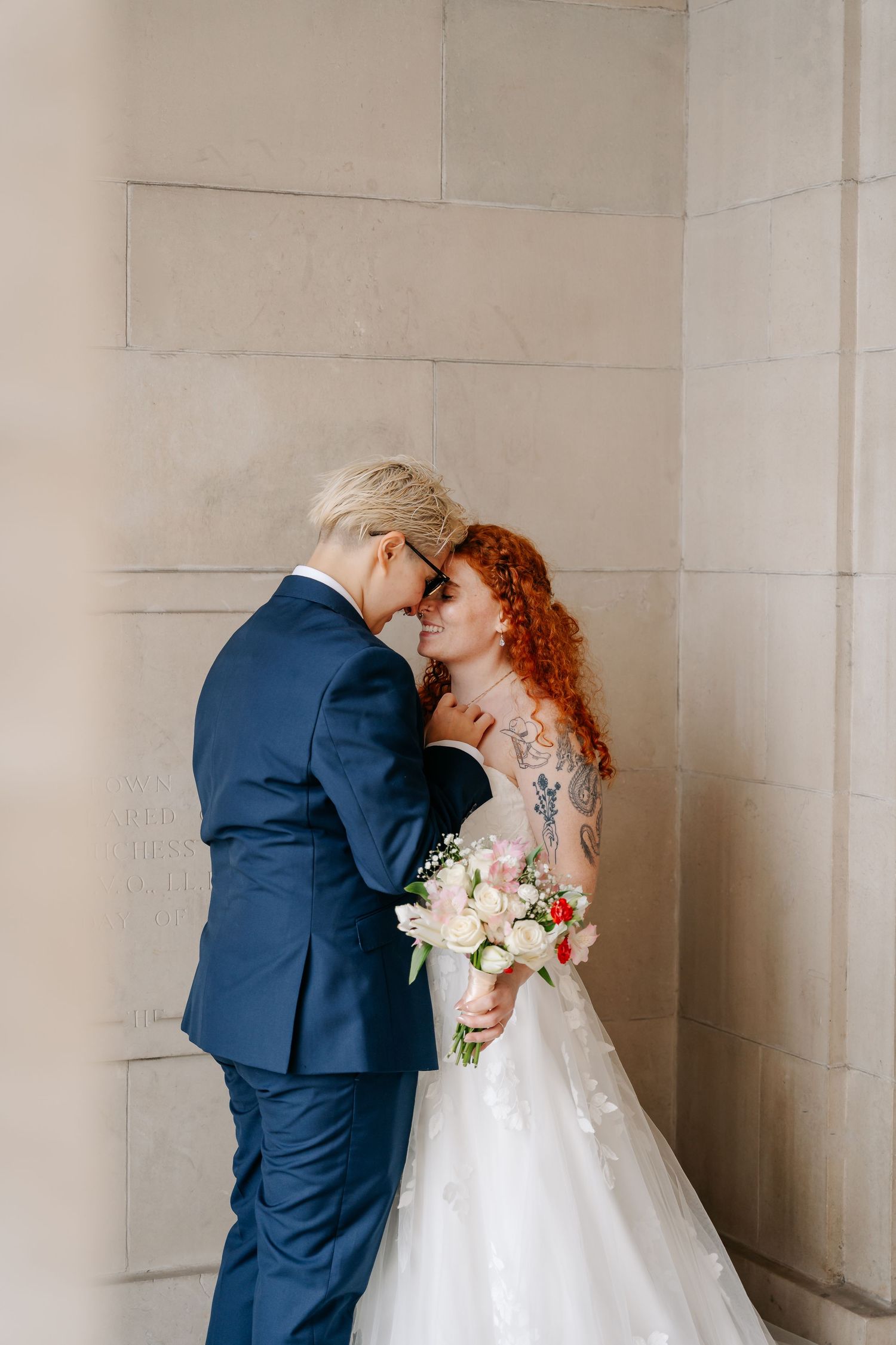 Two brides share a romantic moment, one in a navy suit and one in a white gown holding a bouquet, against a stone wall.
