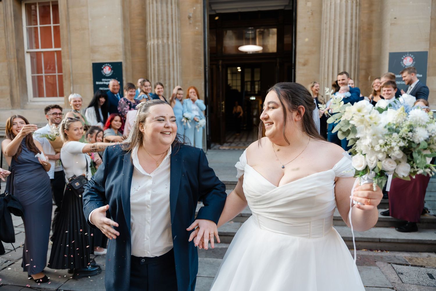 Two brides exit a historic building smiling, one in a white gown holding flowers, guests celebrating behind them.