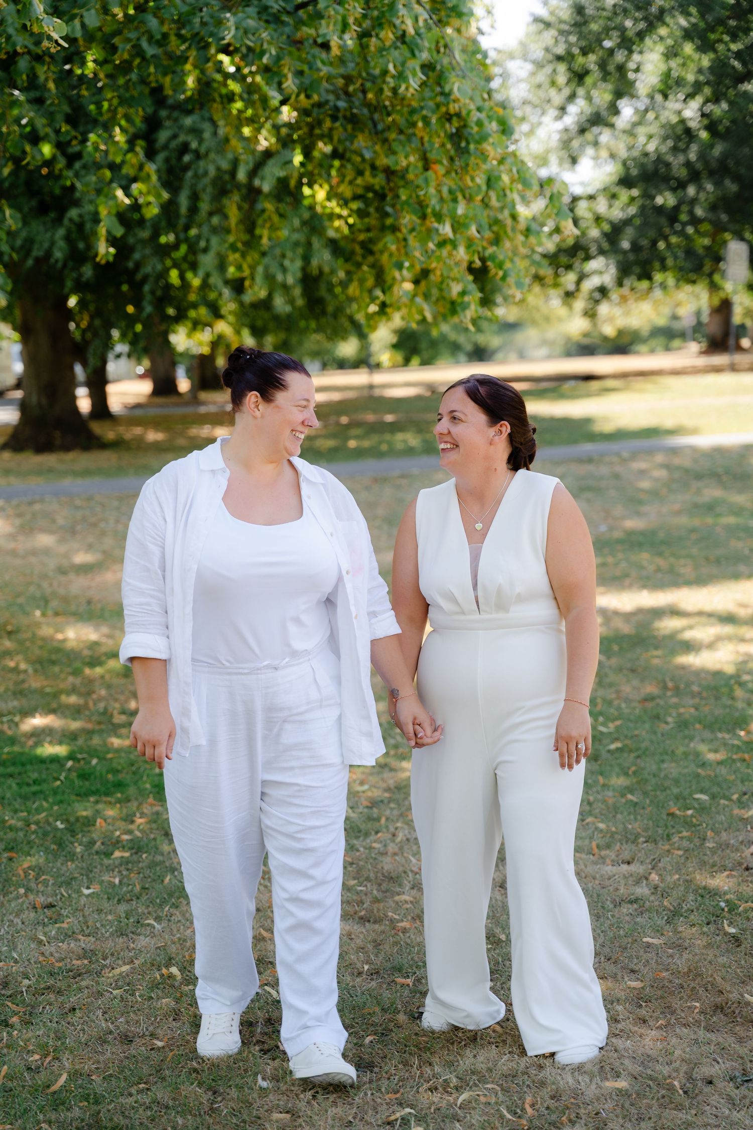 Two women in white outfits hold hands and smile at each other while walking in a sunny park.
