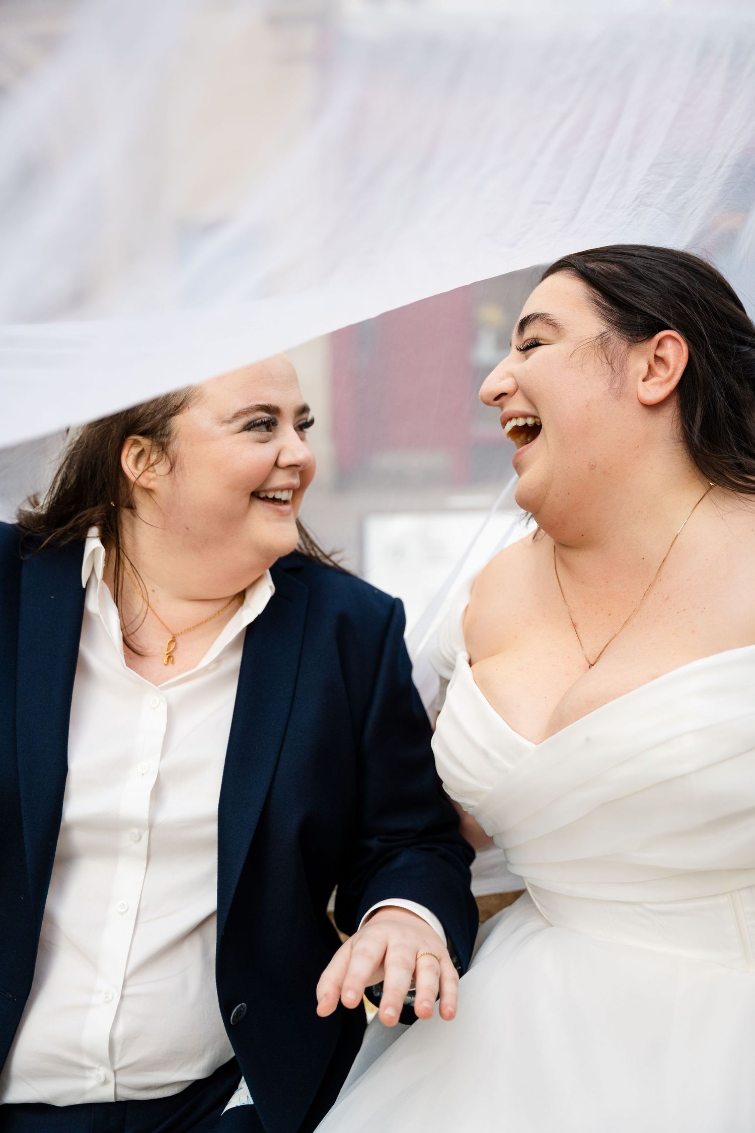 Two brides laughing joyfully under a veil, holding hands, one in a navy suit and one in a white wedding dress.