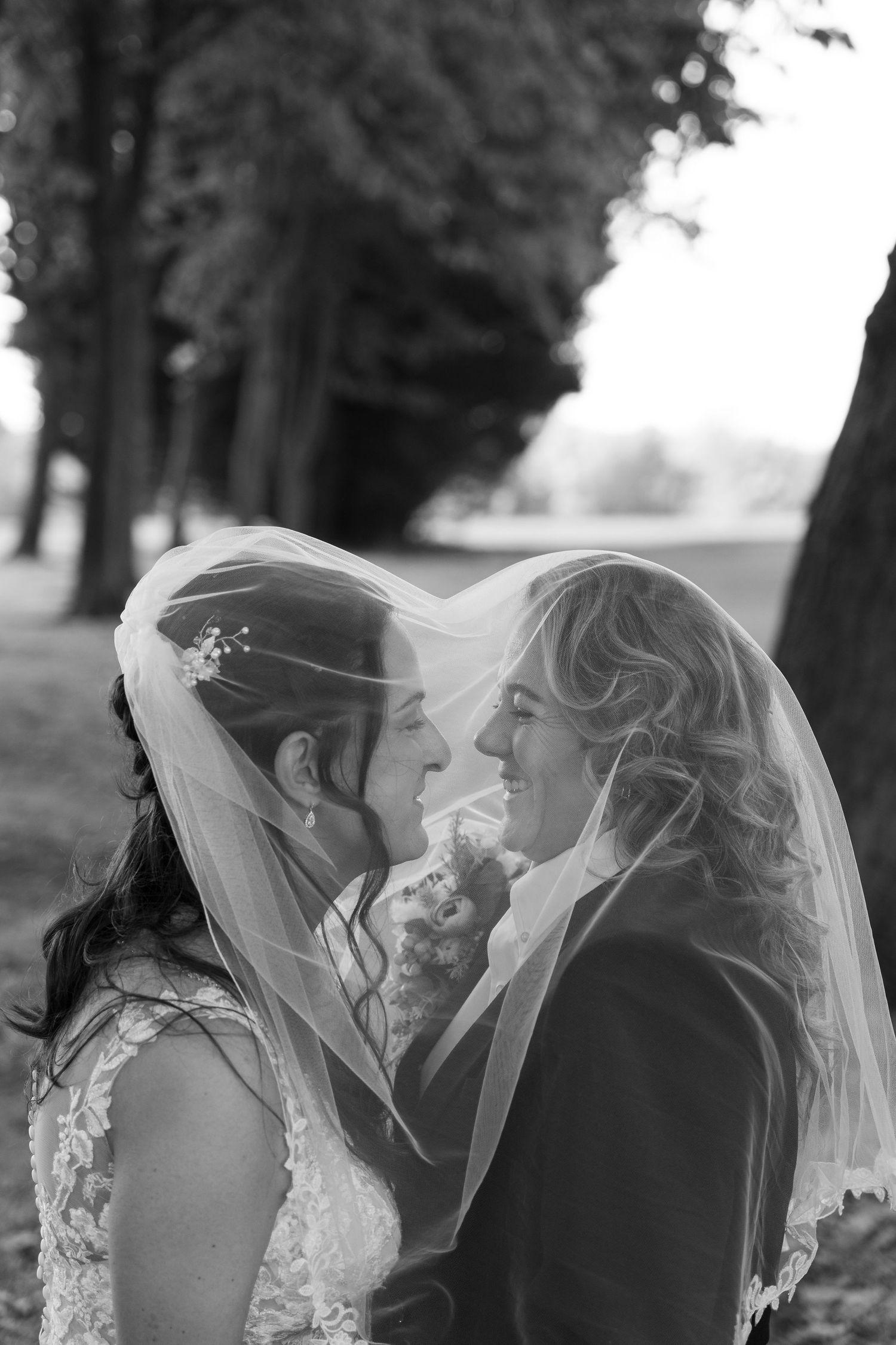 Two brides share a romantic moment under a wedding veil in a black and white outdoor portrait.