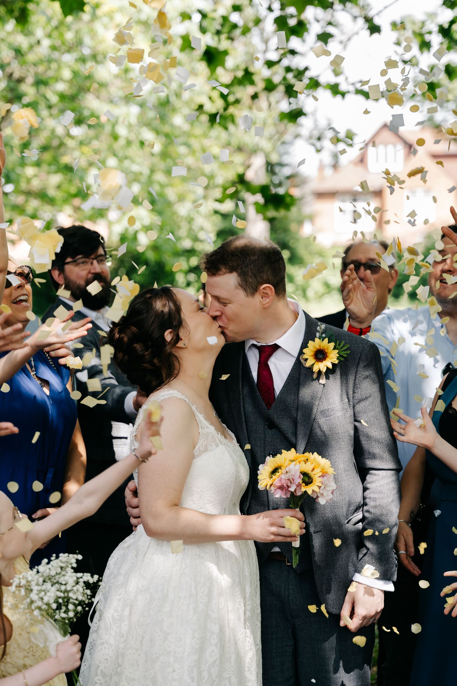 Bride and groom kissing during confetti shower, surrounded by guests outdoors, bride holding sunflower bouquet in white gown.