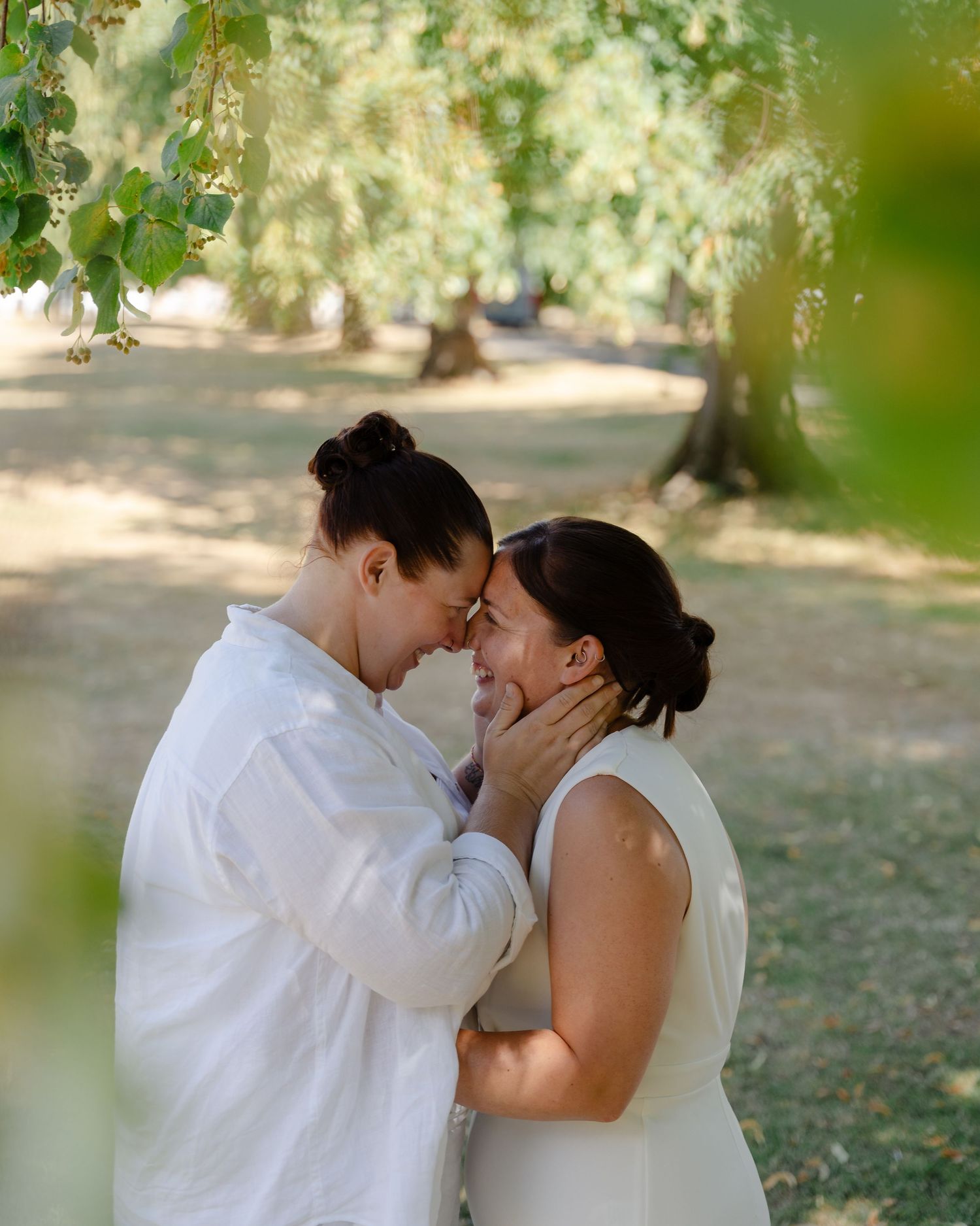 Two brides in white share a tender forehead touch outdoors under lush green trees on their wedding day.