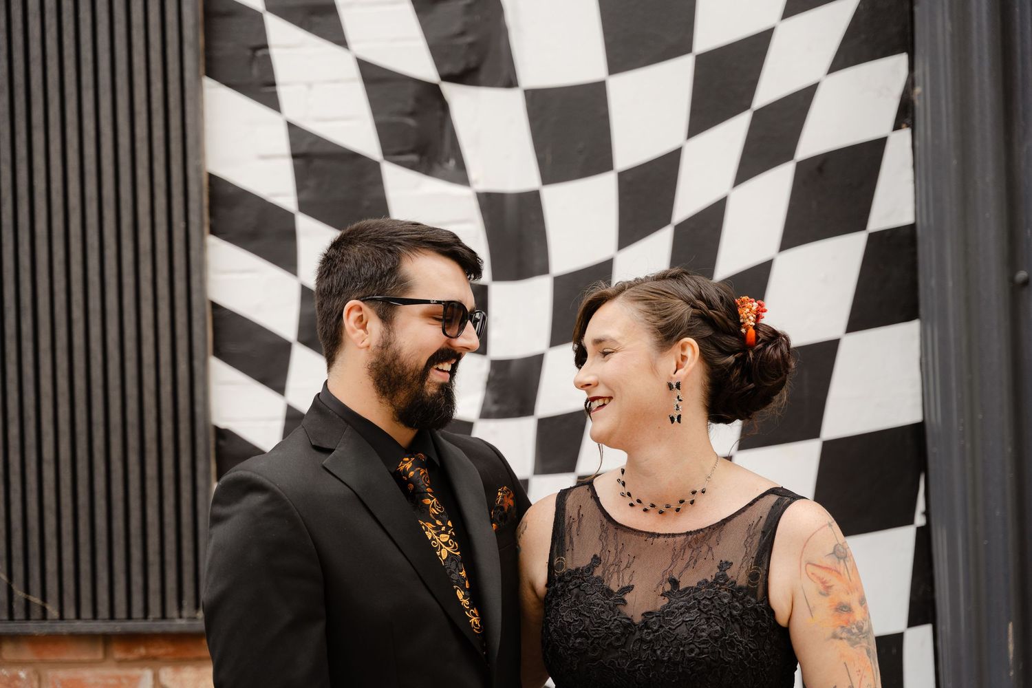 Couple smiling at each other in front of black and white checkered mural, wearing elegant black outfits.