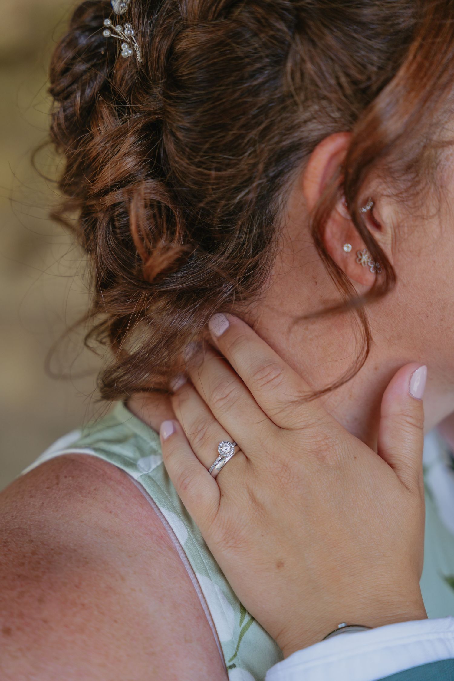 Close-up of bride with updo hairstyle, diamond engagement ring on hand touching neck, small ear piercing visible.