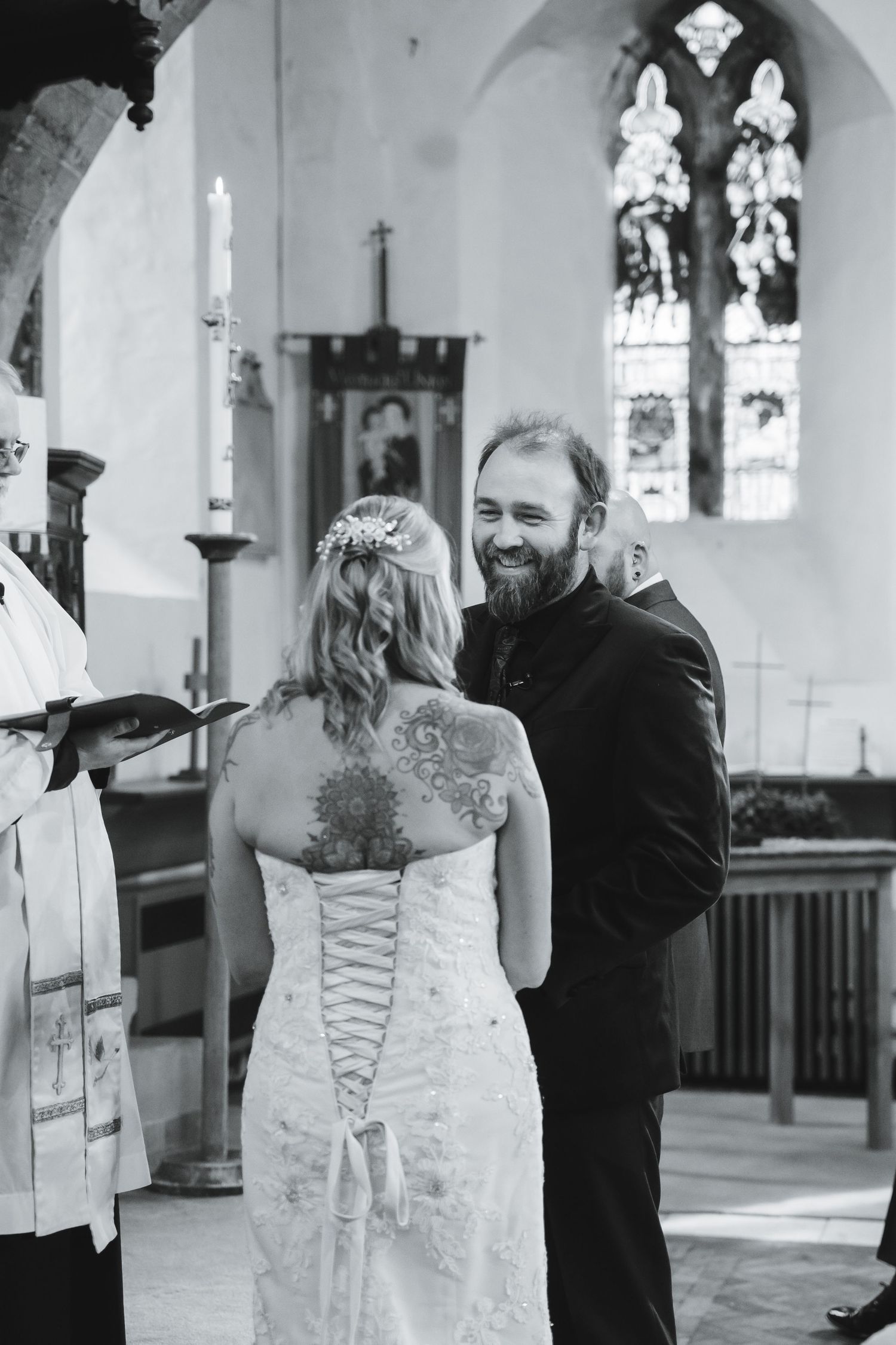 Black and white photo of a bearded groom smiling at tattooed bride during church wedding ceremony.