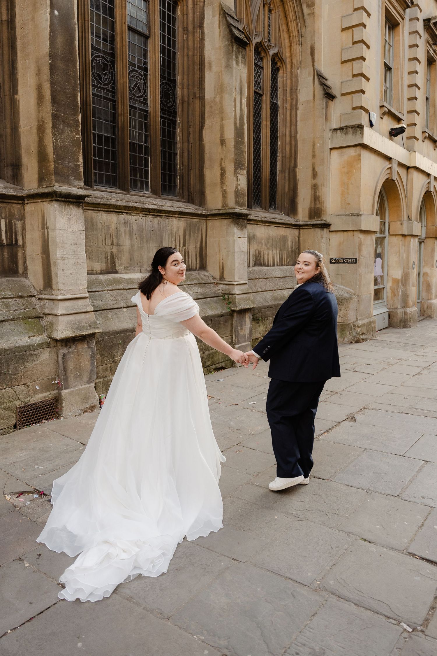 Two brides hold hands outside a historic stone building, one in a white gown and one in a navy suit.
