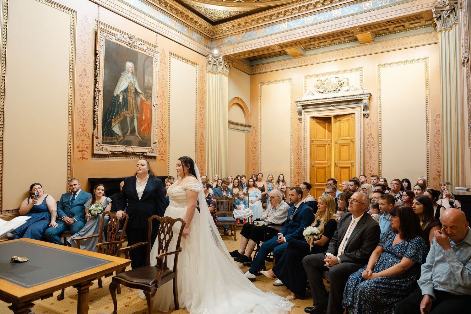 Bride and groom stand at the altar in an elegant ornate hall filled with seated wedding guests during a formal ceremony.