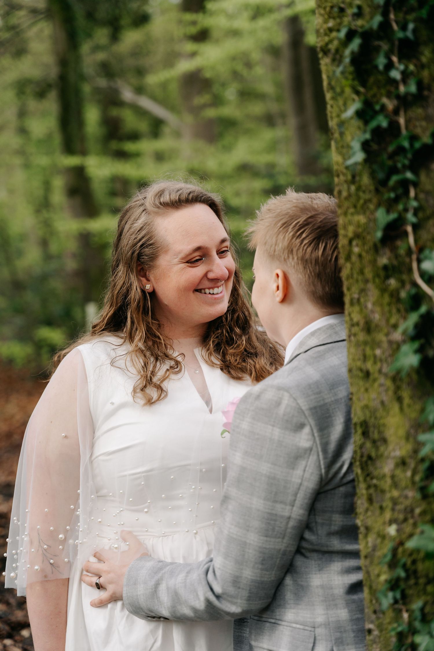 Two brides share a joyful moment in a lush forest, one in a white gown with veil, the other in a grey suit.