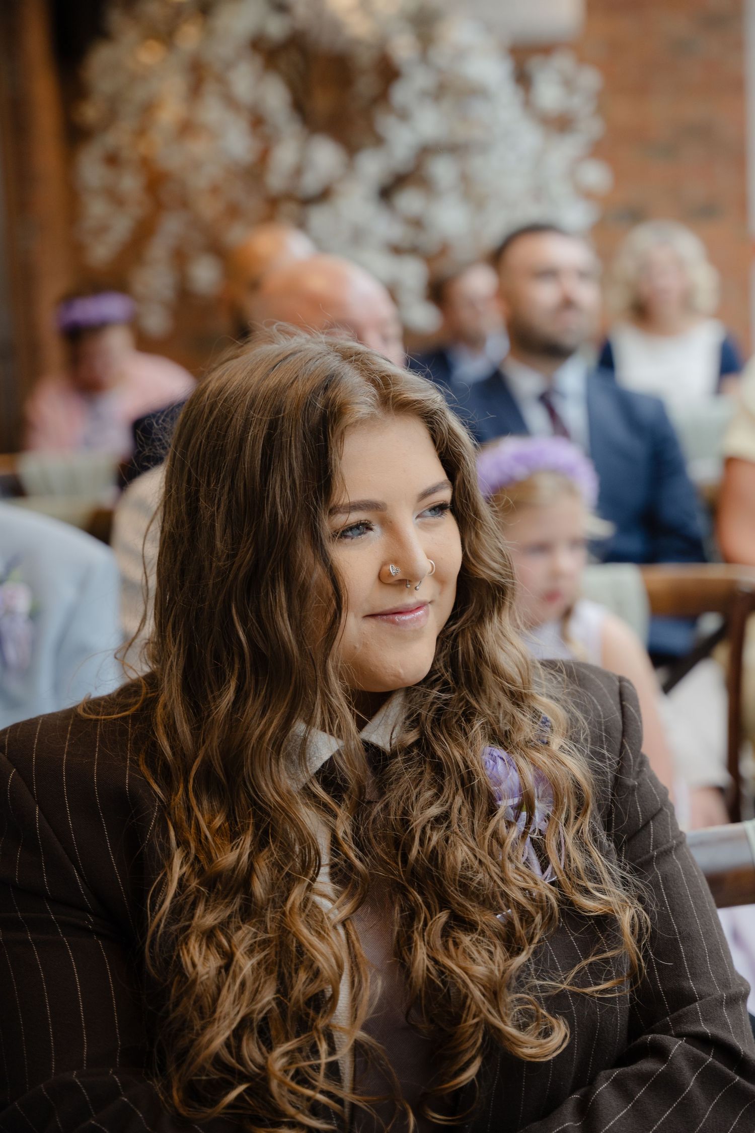 A young woman with long curly brown hair smiles softly at a wedding ceremony, wearing a grey blazer with a floral pin.